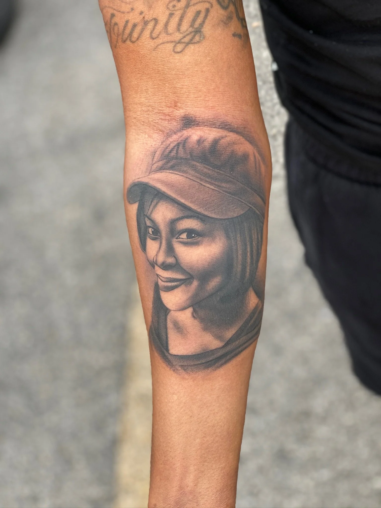 Black and gray realistic portrait tattoo of a woman with a cap on the inner forearm.