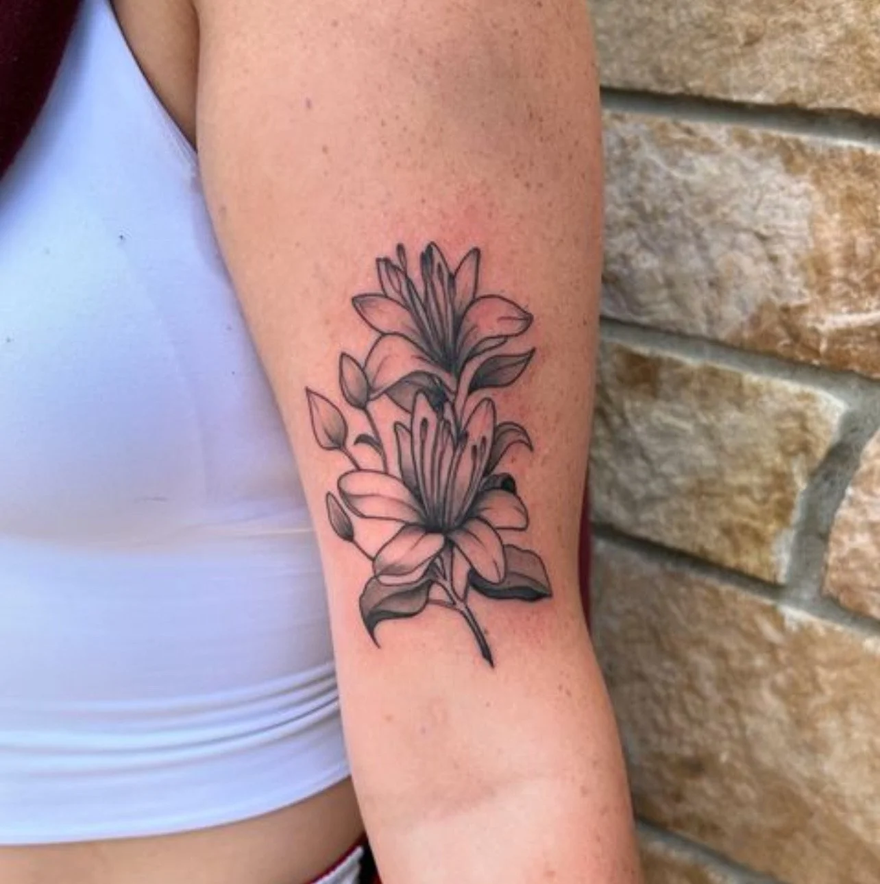 Line drawing of two flowers with leaves on a person's arm, near the shoulder, against a brick wall background.