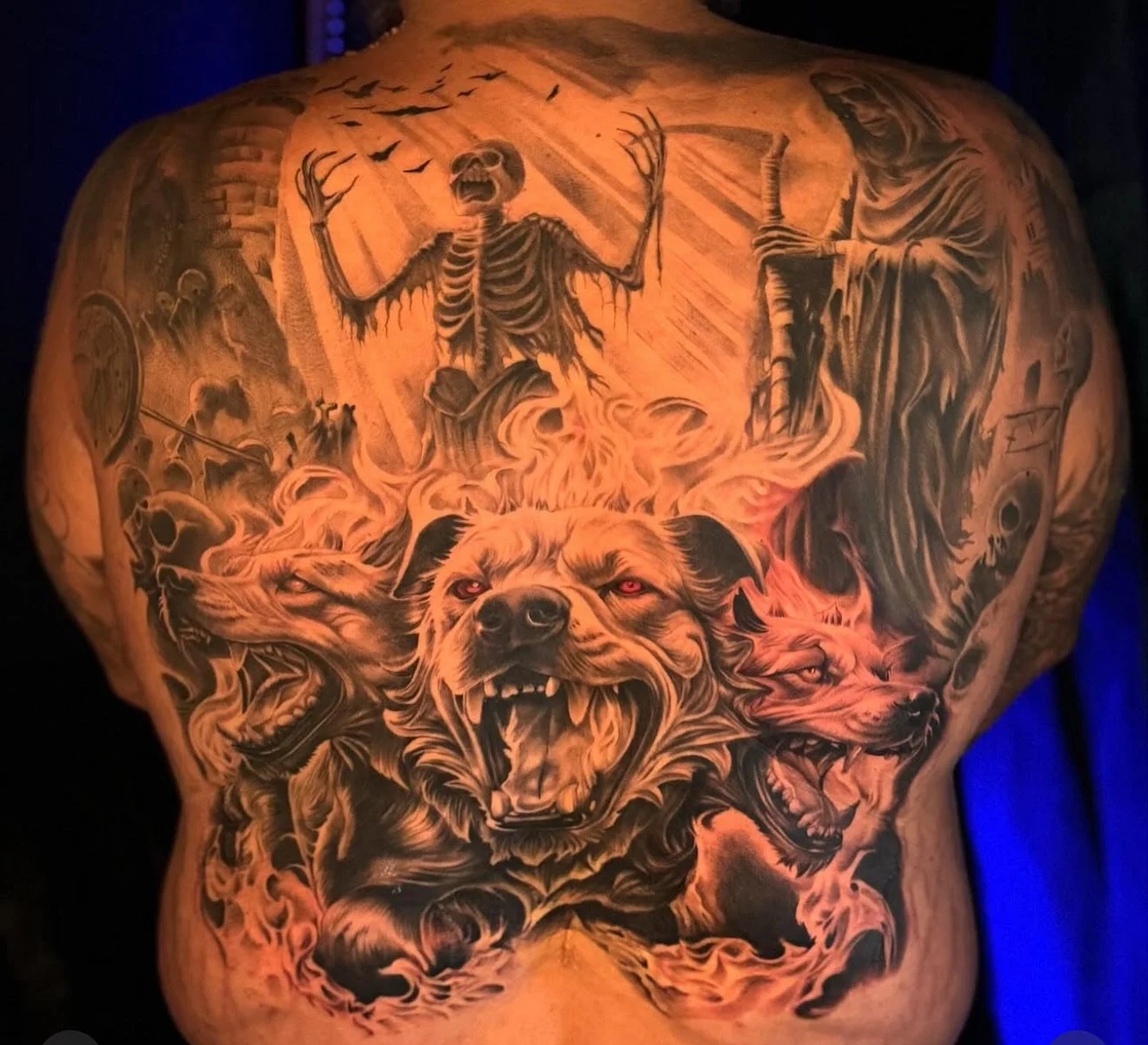 A detailed black and gray tattoo on a person's back depicting a scene with a fierce, three-headed dog with glowing eyes, surrounded by flames. Above the dog, there is a skeleton with outstretched arms and an open mouth, and a robed figure holding a s