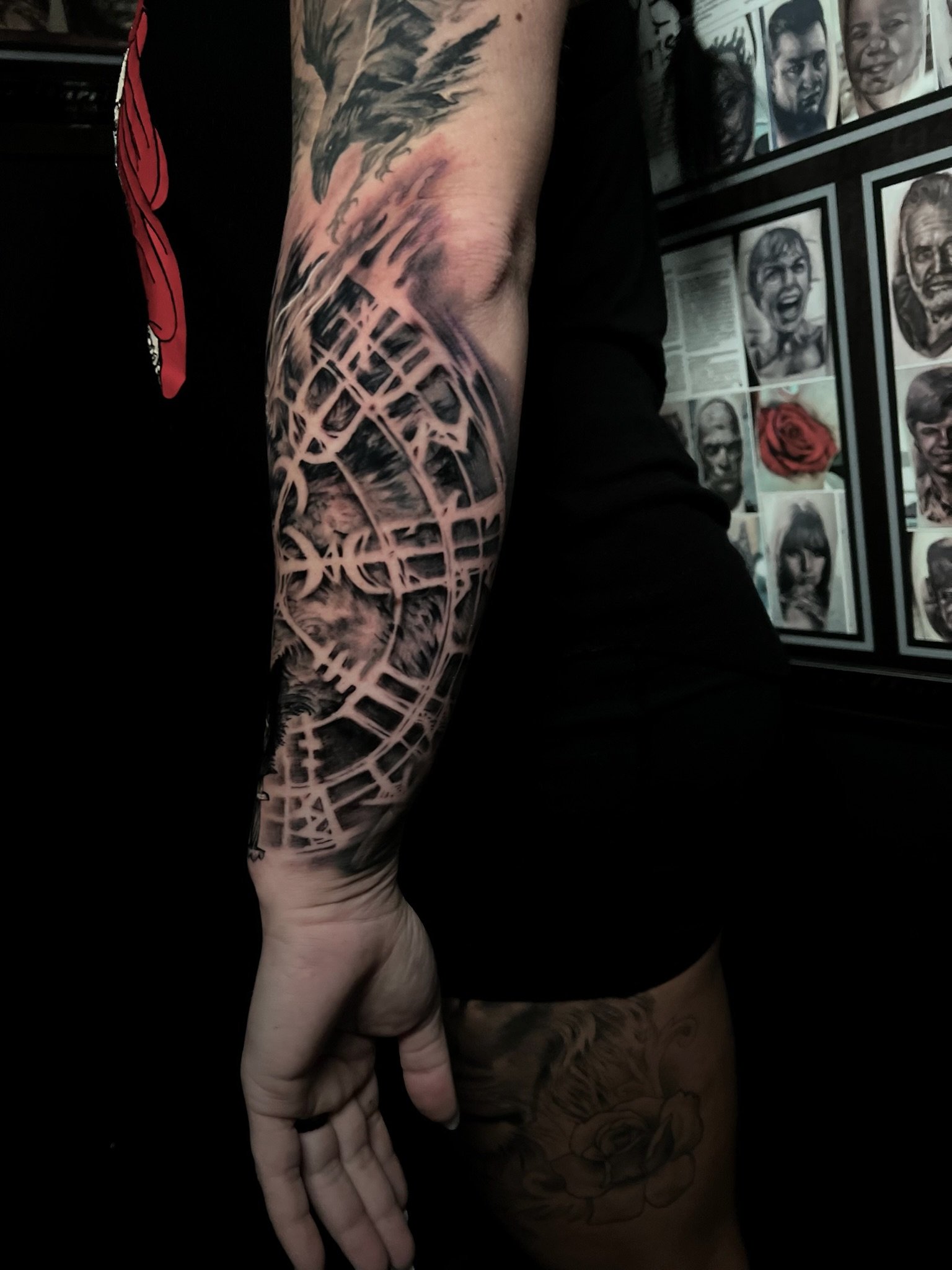 Close-up of a person's arm with a detailed black and gray tattoo of a globe and abstract lines, with additional tattoos on the hand and leg, standing in front of framed artwork and photographs.