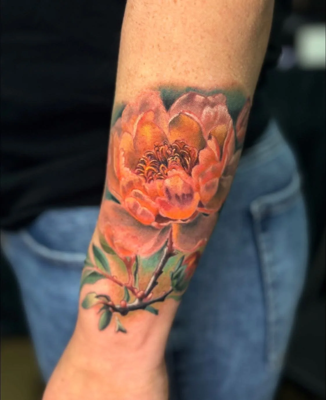 Colorful realistic flower tattoo in orange, yellow, and pink shades on a person's forearm, with green leaves and a branch.