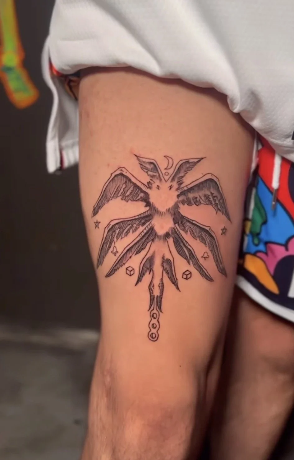 Tattoo of a dragonfly on a person's thigh with intricate line details, surrounded by small celestial and geometric symbols.