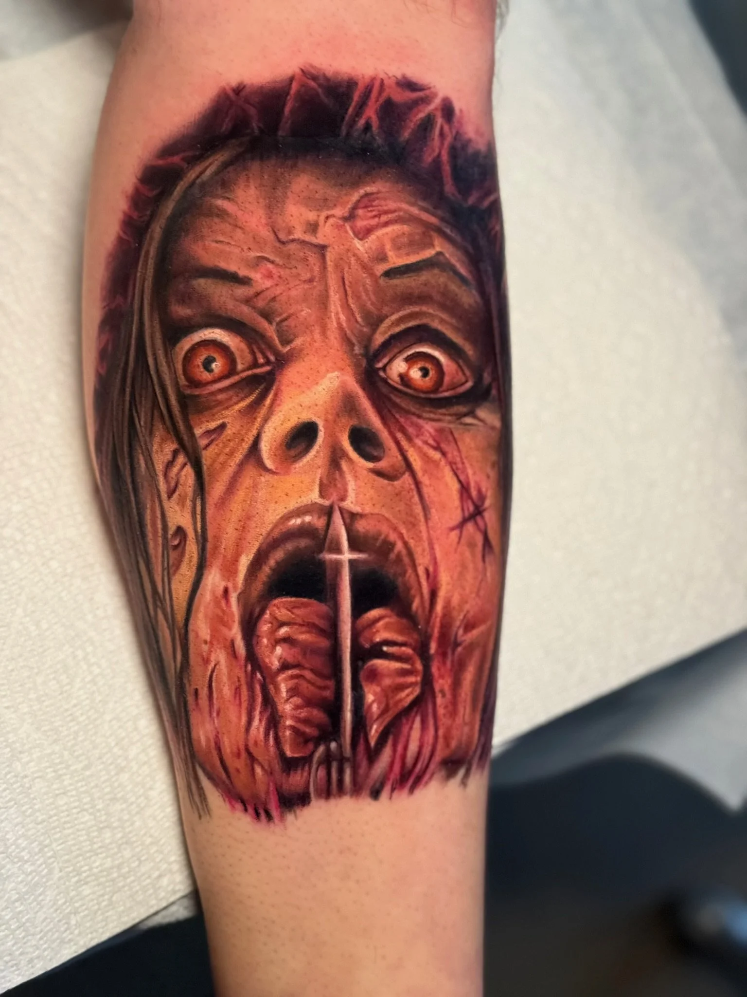 Tattoo of a horror-themed face with wide-open eyes, an open mouth with tongue out, and detailed shading in fiery tones.