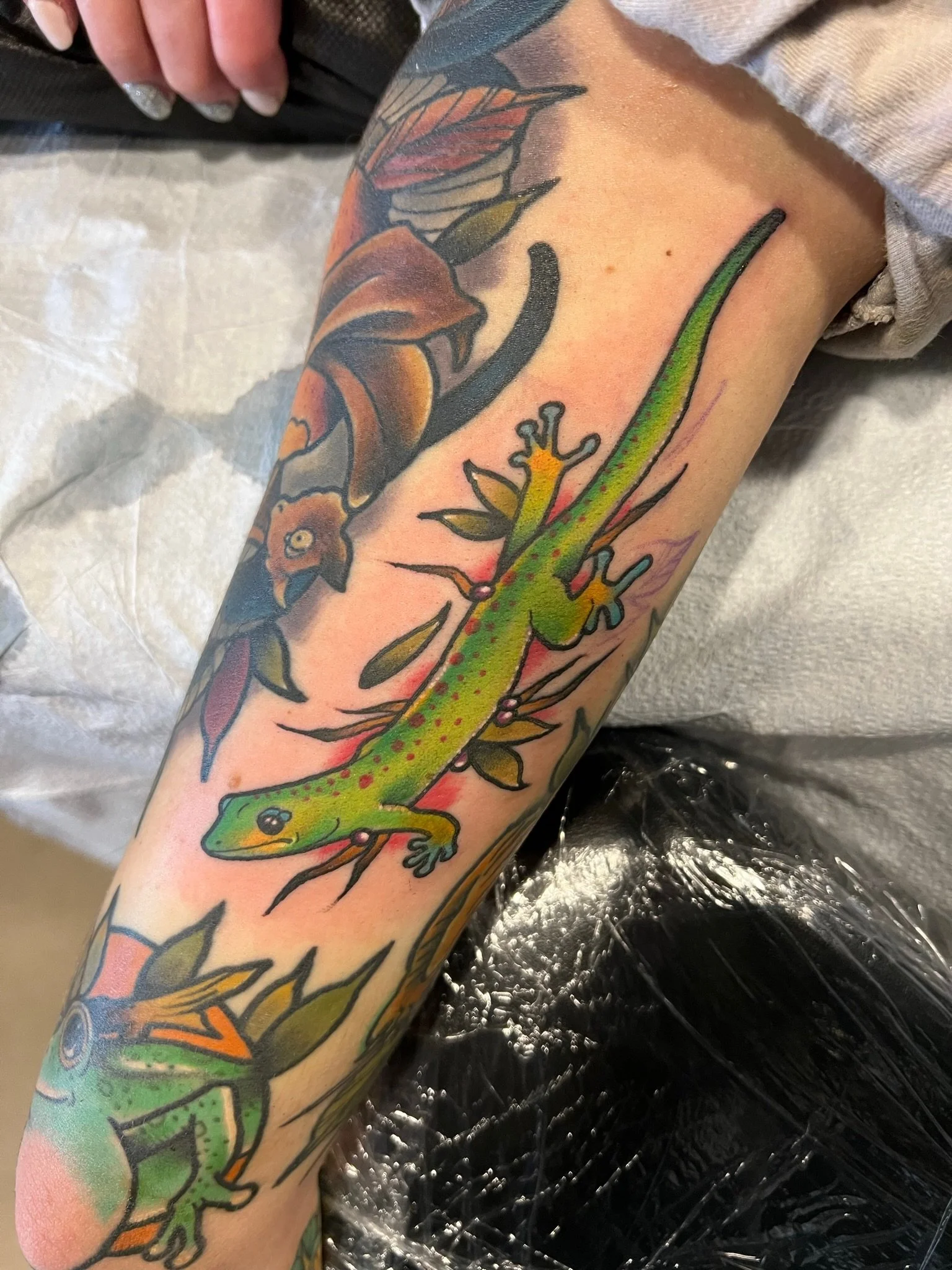 Tattoo of a green gecko with red spots climbing a branch, with a floral design of orange and red leaves in the background.