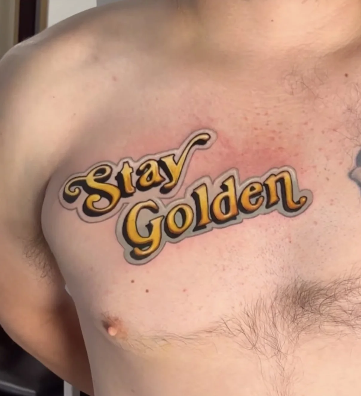A person's chest with a tattoo that says "Stay Golden" in stylized yellow and black lettering.