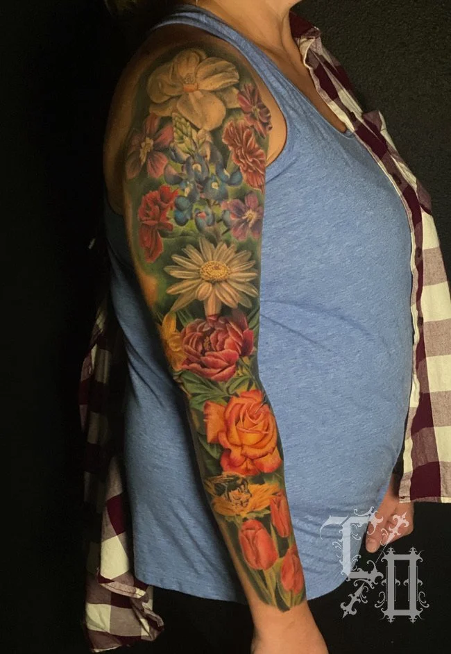 Colorful floral tattoo sleeve on woman's arm, featuring large yellow, white, and orange flowers with green leaves.