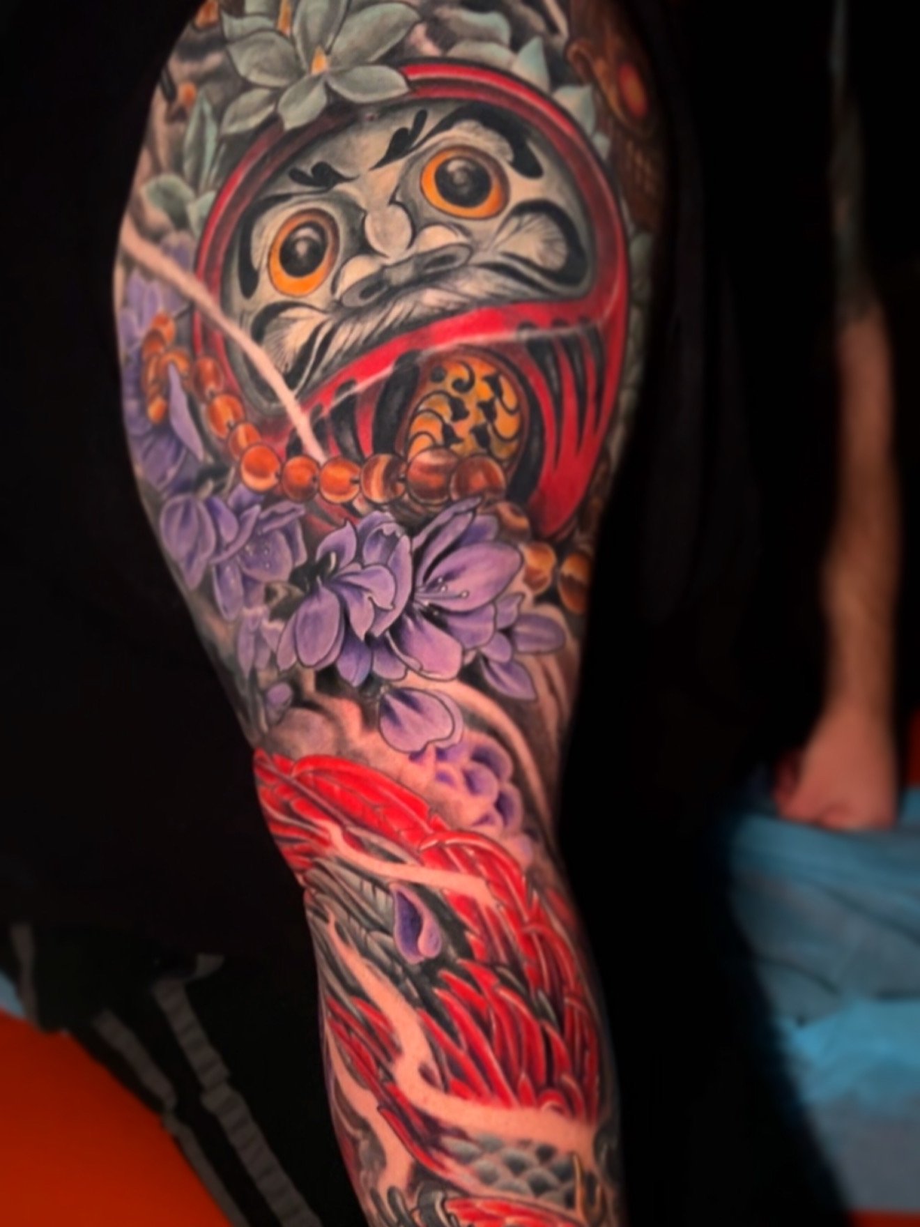 Colorful tattoo on a person's arm featuring a traditional Japanese Daruma doll, purple flowers, red and black swirling waves, and intricate details.