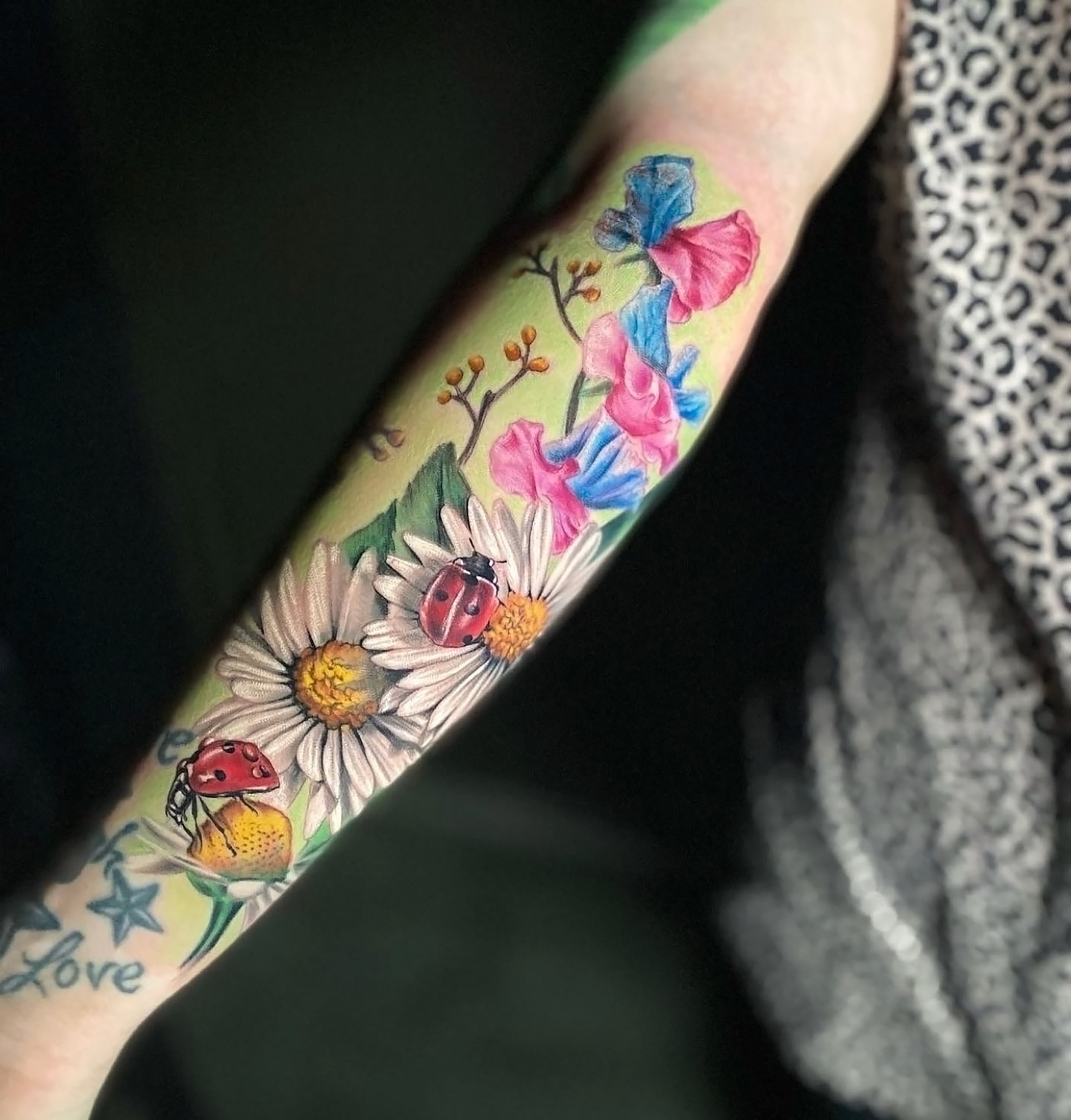 Close-up of a tattoo on a person's arm featuring colorful flowers, a ladybug, and a bee, with partially visible text.