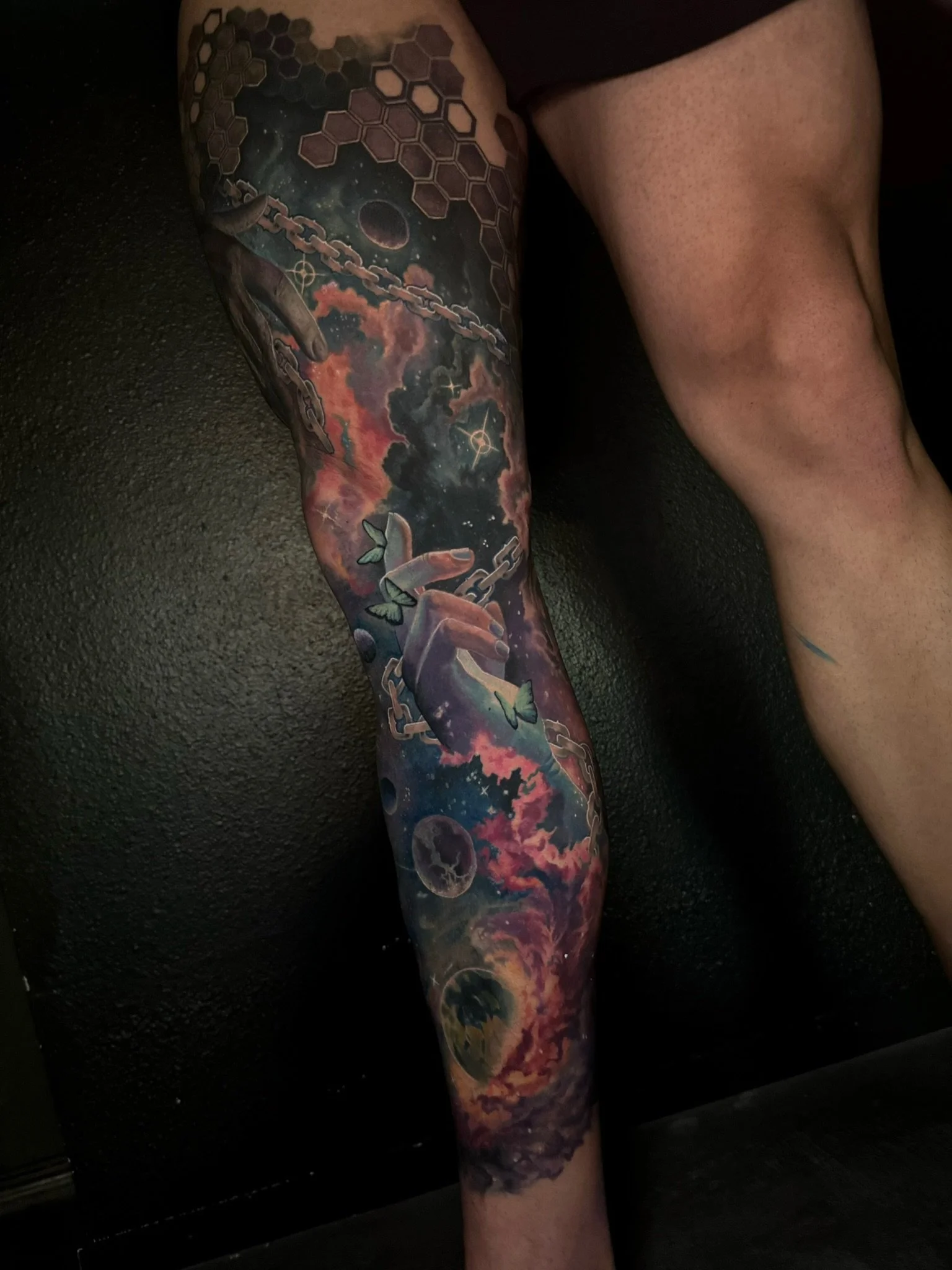 A colorful full sleeve tattoo featuring cosmic elements such as planets, moons, stars, clouds, and geometric shapes on a person's arm.