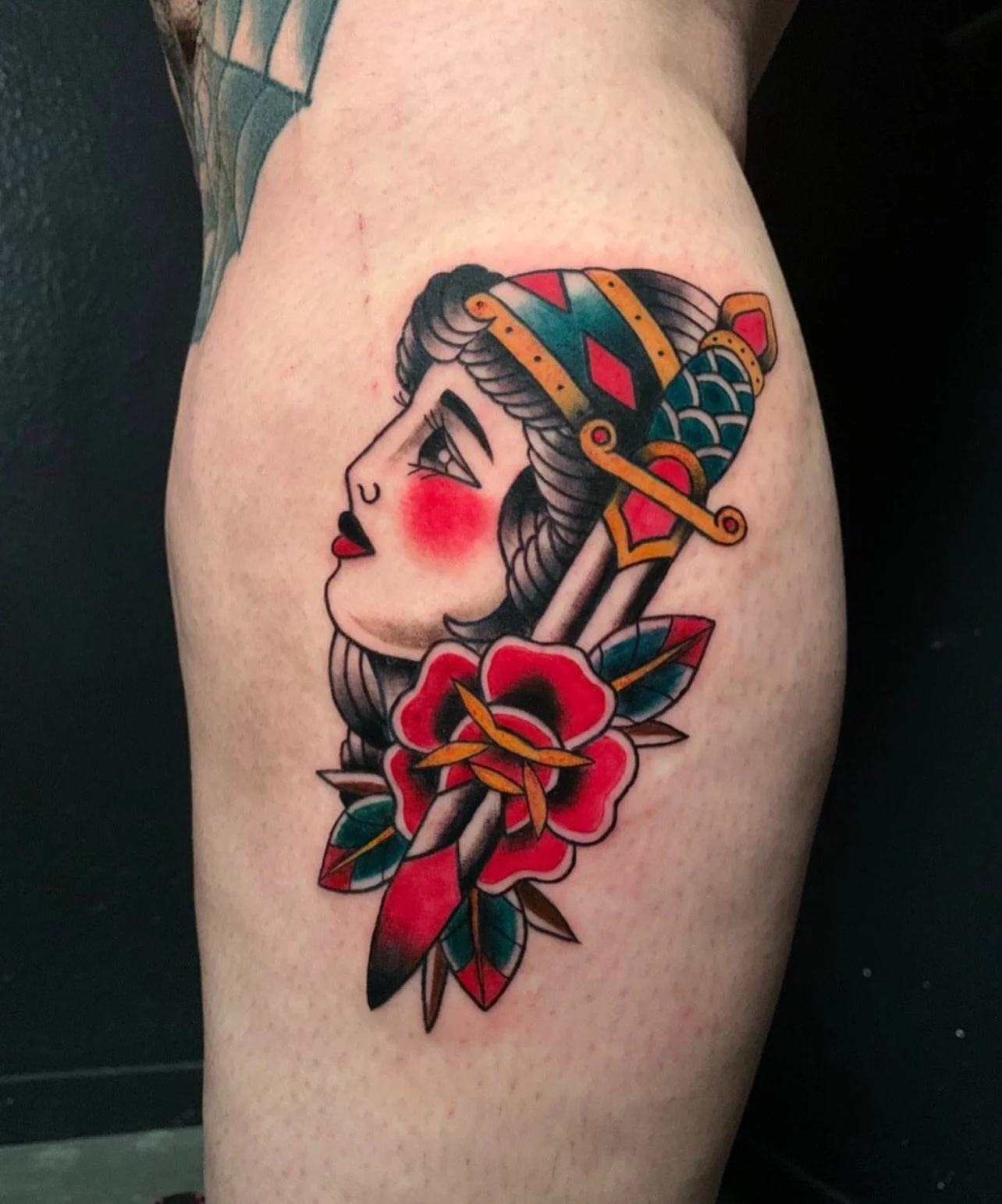 Tattoo of a woman’s face with traditional tattoo style, red cheeks, black hair, and a decorative headpiece, with a red flower and leaves beneath.