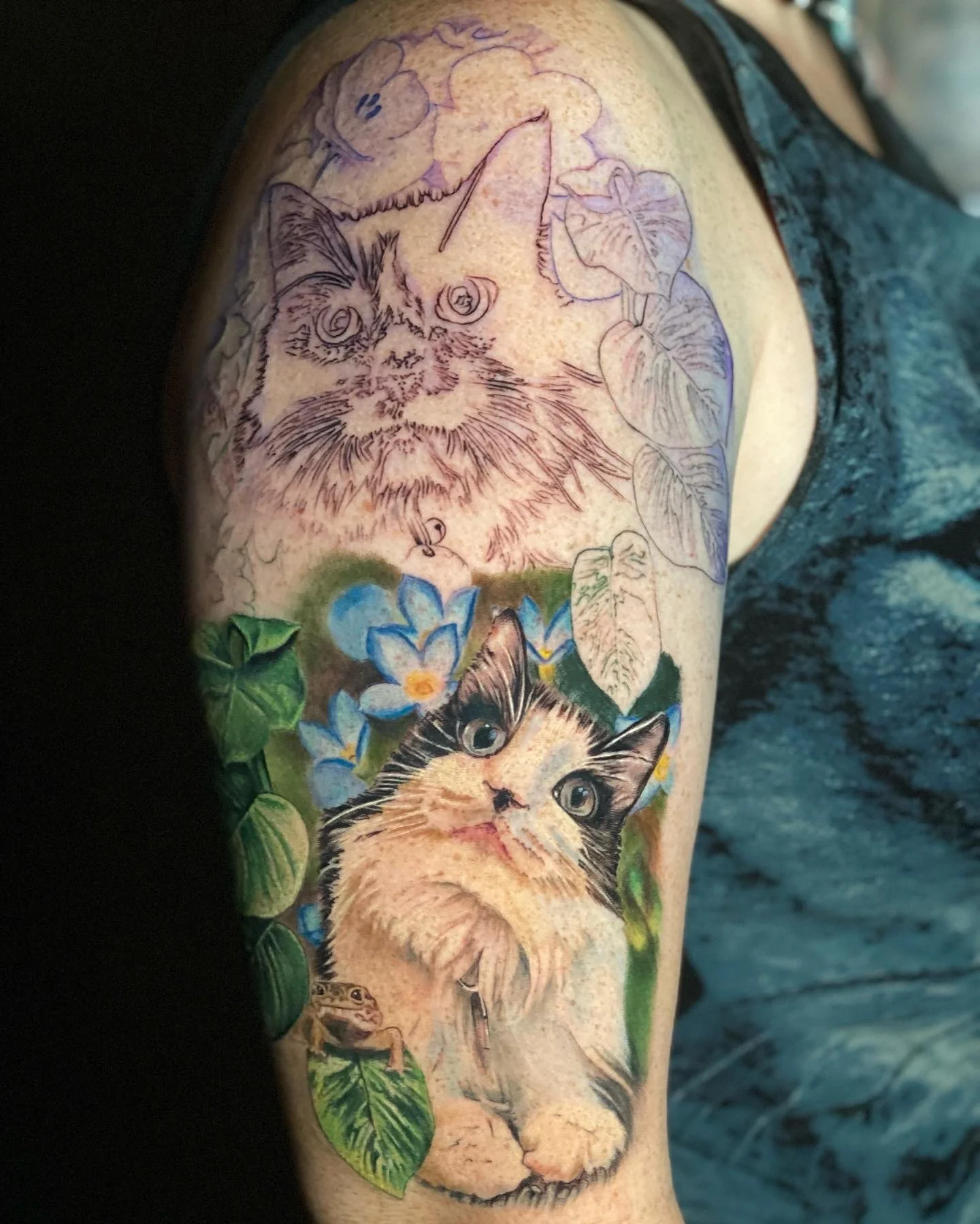 Tattoo of a cat surrounded by blue flowers and green leaves, with a sketch of a kitten and a ladybug on the skin.