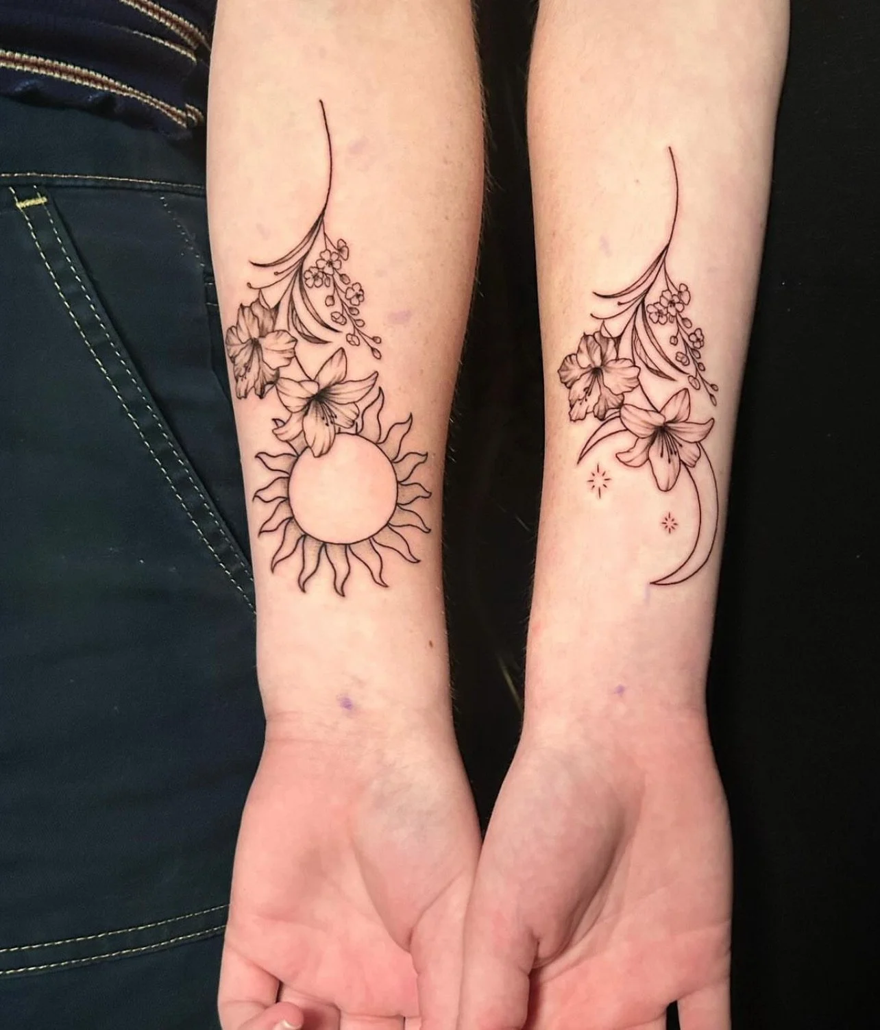 Two forearms with matching black ink floral tattoos, featuring lilies, a sun, moon, stars, and swirling lines.