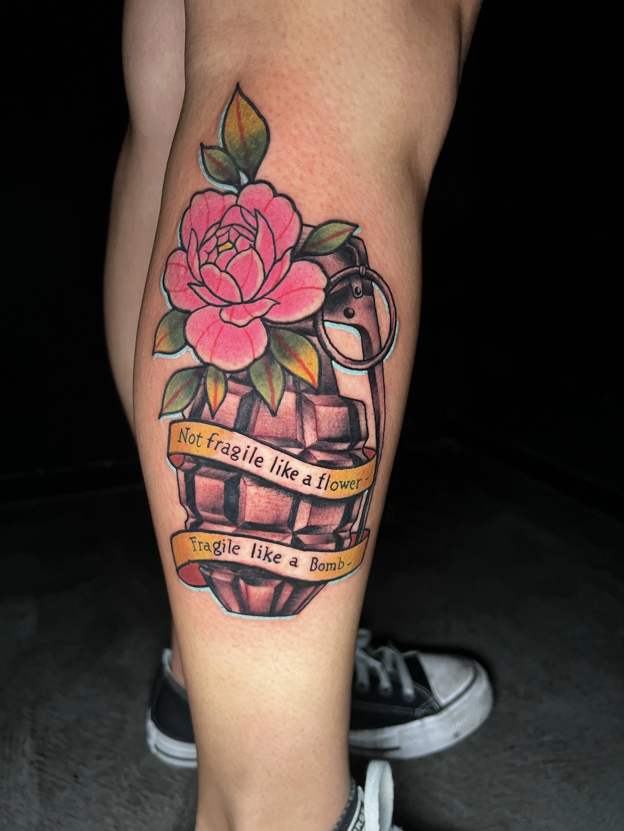 tattoo of a colorful grenade with a pink flower and green leaves, with a banner that says "not fragile like a flower. fragile like a grenade" on a woman's outer calf