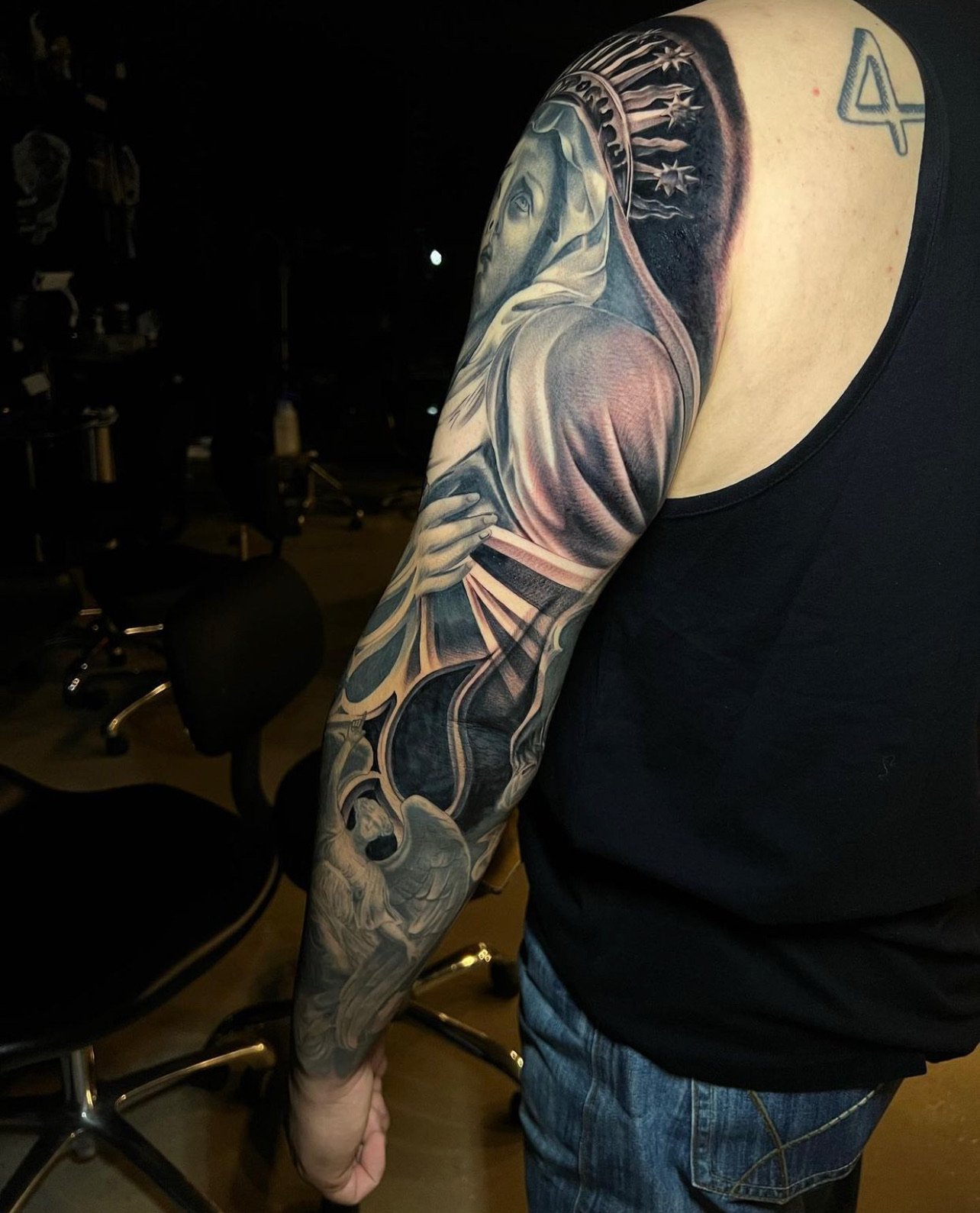 Person with a detailed black and gray tattoo sleeve depicting a religious scene, including a woman and a skull, on their arm.
