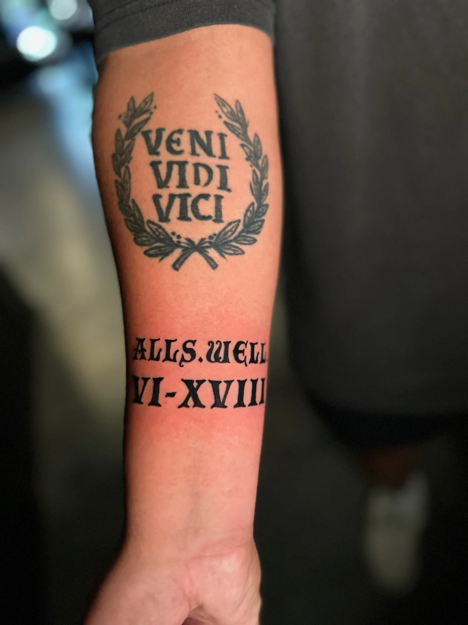 Tattoo on arm with Latin phrase "Veni Vidi Vici" inside a laurel wreath and another phrase in Roman numerals "X-VIII-M-XX" that reads "All's well".