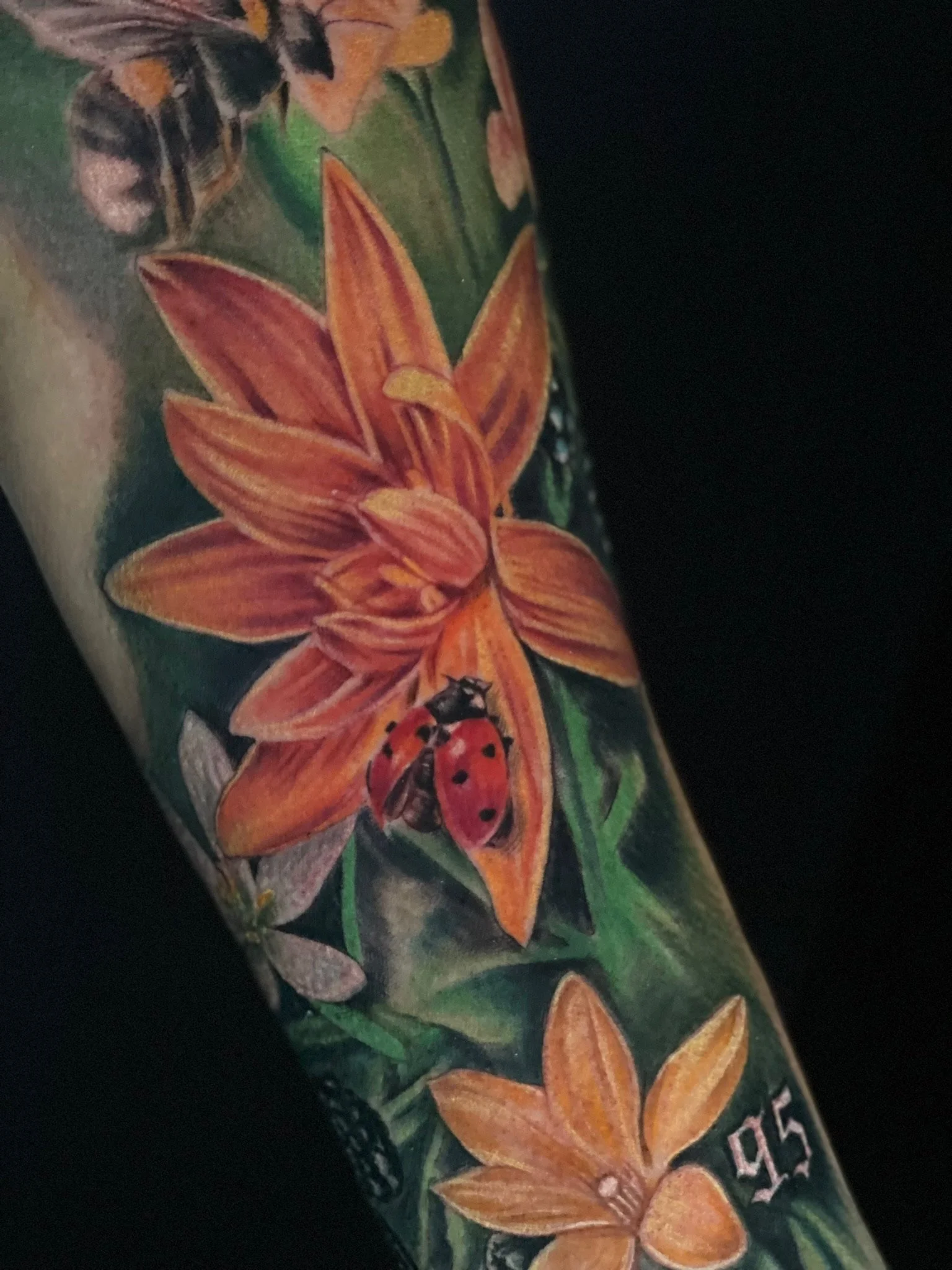 Close-up of a colorful tattoo of orange lilies, a ladybug, and a bee on someone's arm.