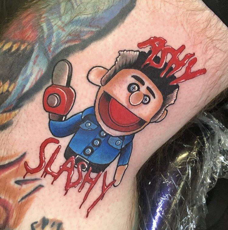 a red, blue, and grey tattoo that says "ashy slashy" written in blood with Ash from the TV show and Evil Dead movie franchises, designed as a hand puppet and holding a chainsaw on a mans inner thigh 