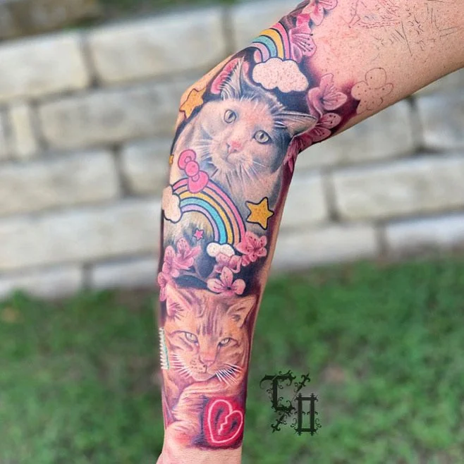 Tattoo of two cats' faces, surrounded by rainbows, clouds, stars, and pink flowers on a person's arm.