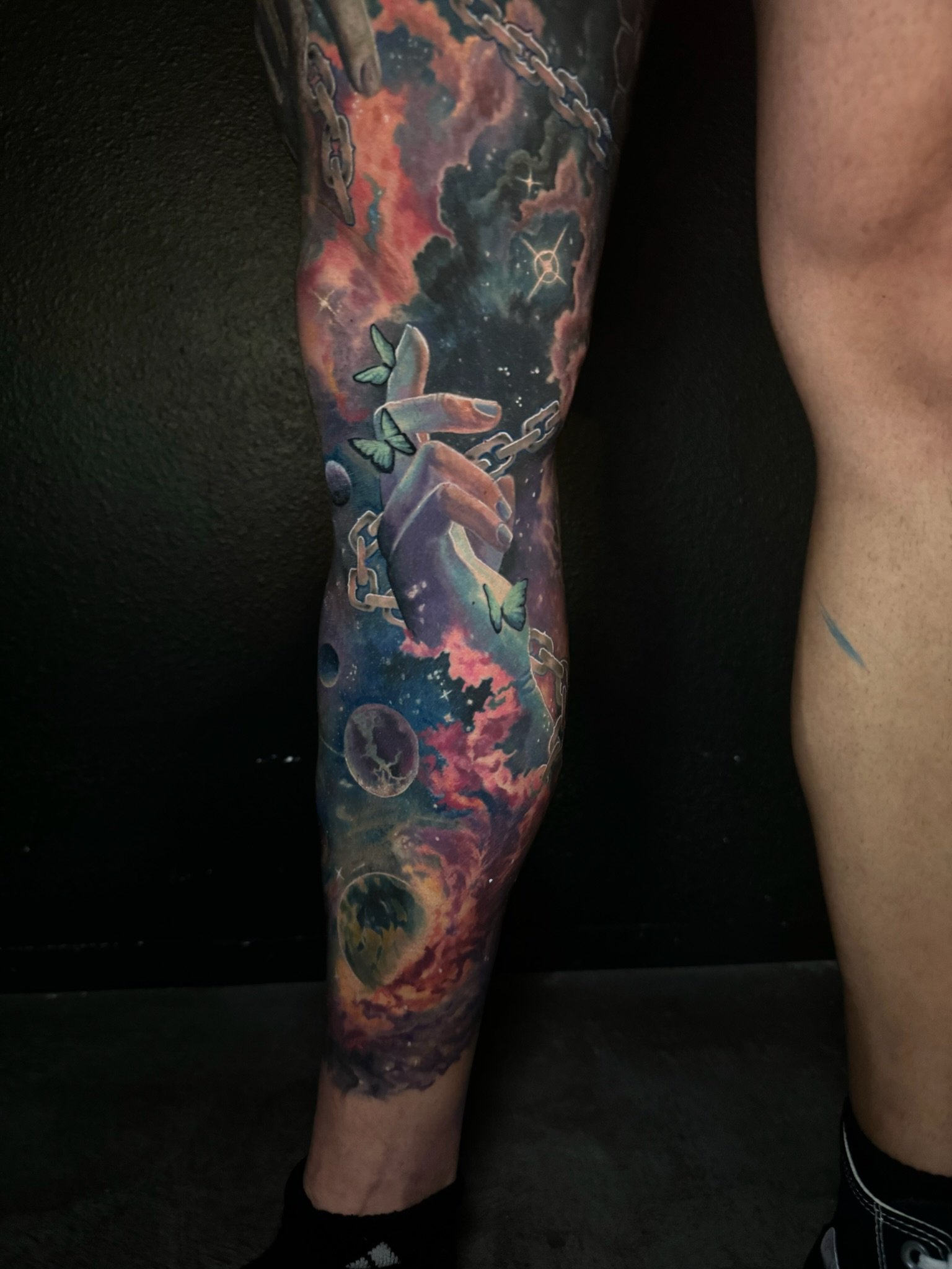 Colorful tattoo sleeve featuring a cosmic scene with planets, a hand holding a chain, butterflies, and a nebula.