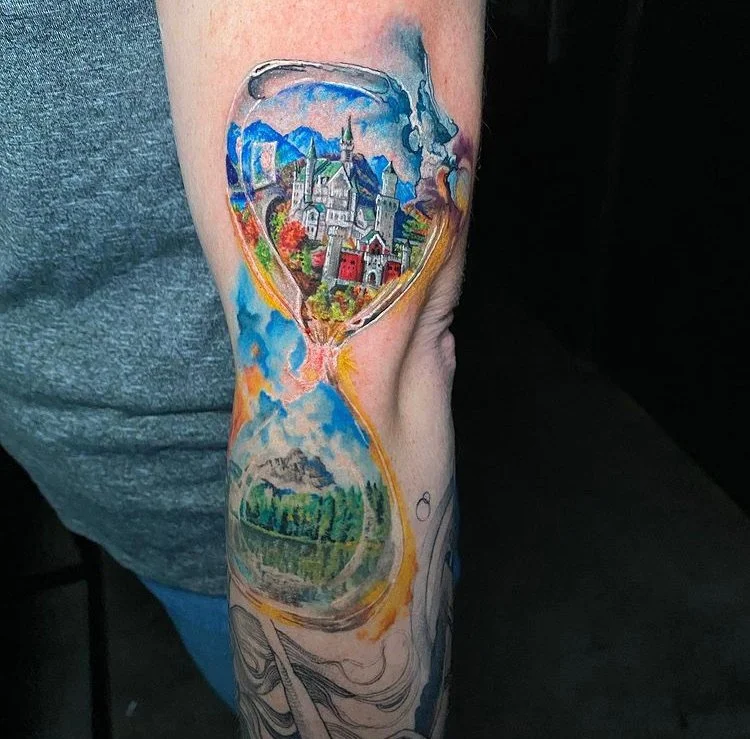 A colorful tattoo of an hourglass on a person's arm, with a detailed cityscape and mountains inside the upper glass, and nature scenery with trees and water inside the lower glass.
