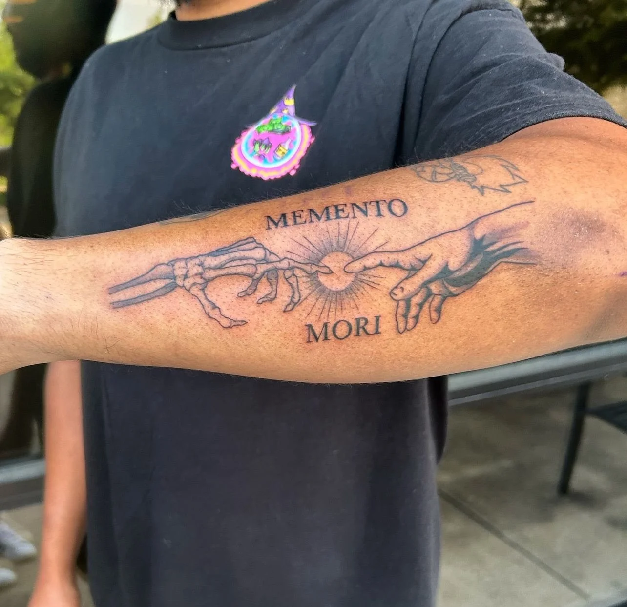 Tattoo of hands reaching towards each other with a sun in the background, with the words 'MEMENTO MORI' above and below.
