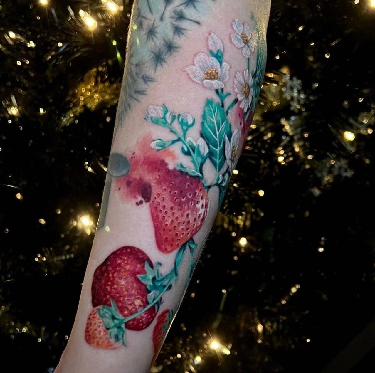 Tattoo of strawberries and white flowers with green leaves on a person's arm, with a background of a decorated Christmas tree with lights.