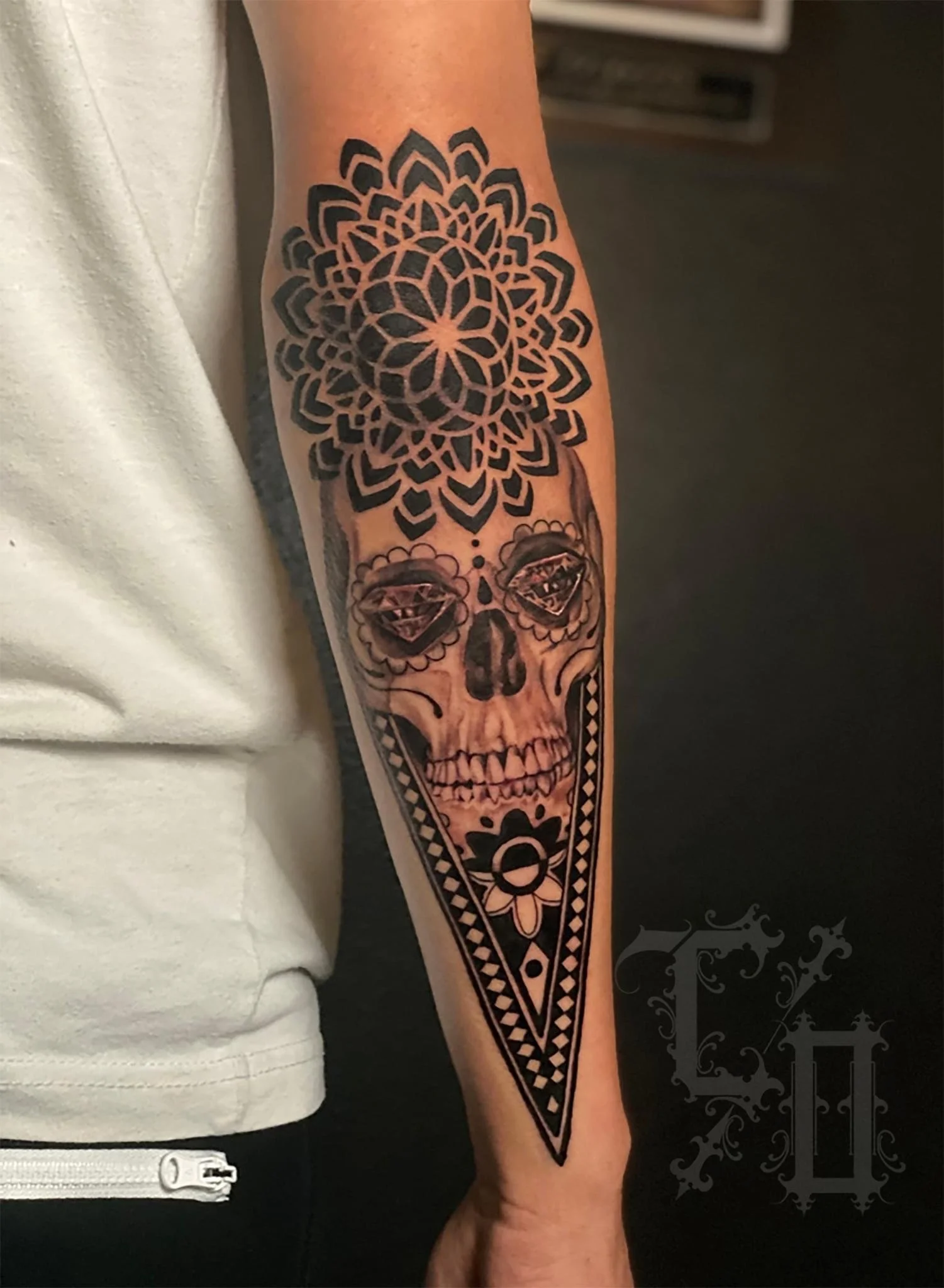 A tattoo on a person's forearm featuring a stylized skull with flower-like details, a large mandala pattern above it, and a triangle with a flower at the wrist.