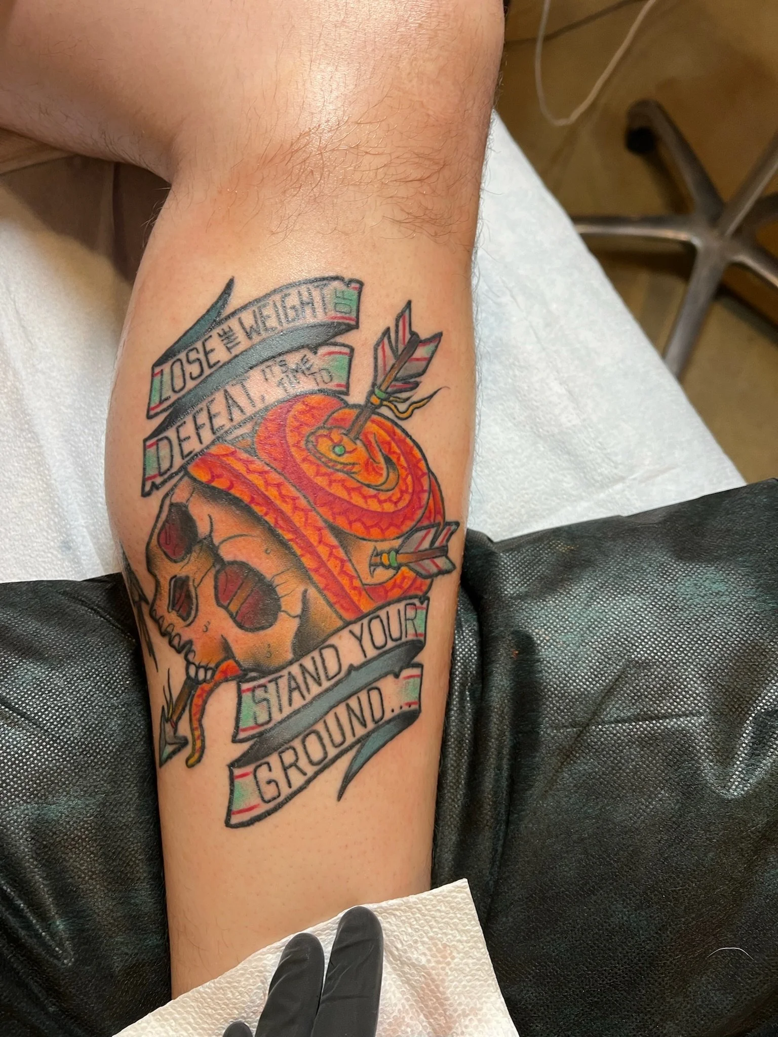 Tattoo of a skull with a snake wrapped around it, an arrow piercing through the snake, and a banner with the words 'Lose the weight, defeat it, stand your ground'.