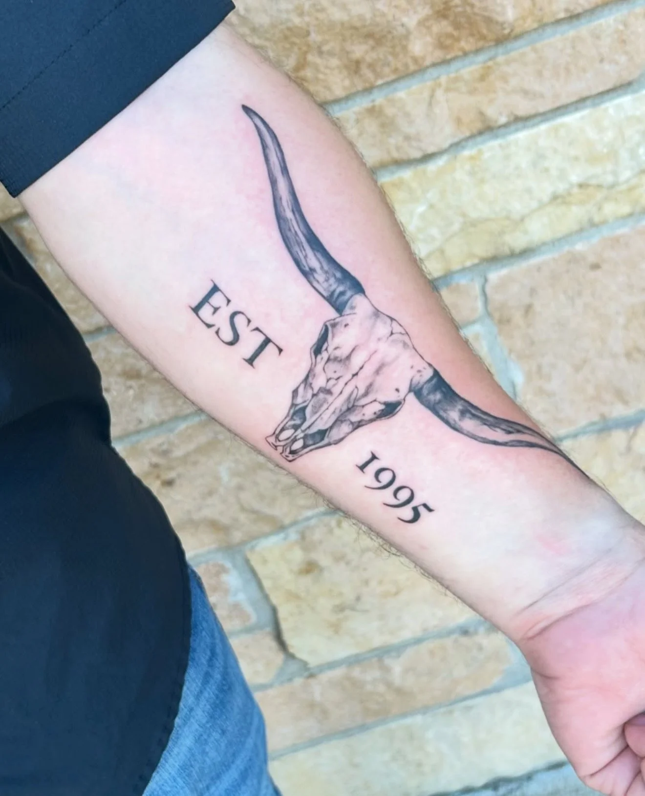 Tattoo of a bull skull with long horns, with the words "EST" and "1995" beside it, on a person's forearm.