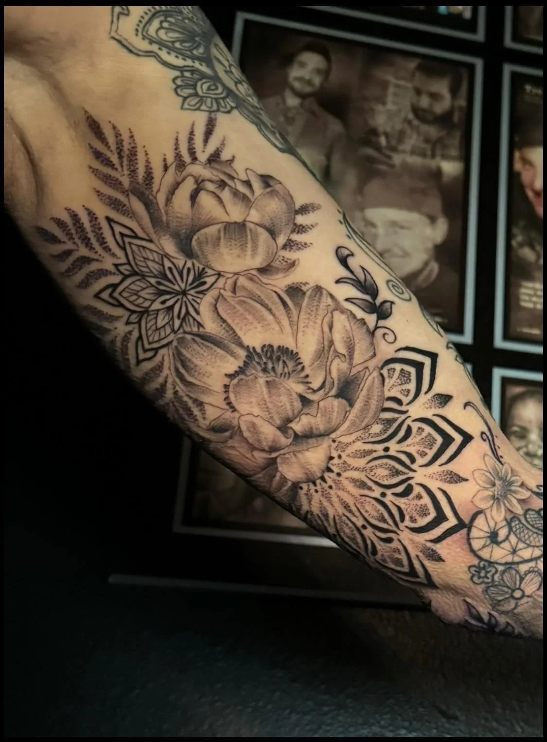 Black and gray floral tattoo sleeve featuring large blooms, leaves, and intricate patterns on a person's arm, with framed vintage-style photographs in the background.
