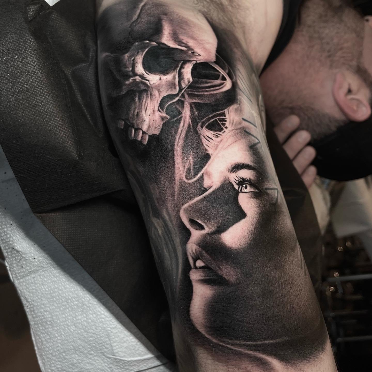 A black and gray tattoo on a person's arm depicting a skull facing a woman's face with an eye visible.
