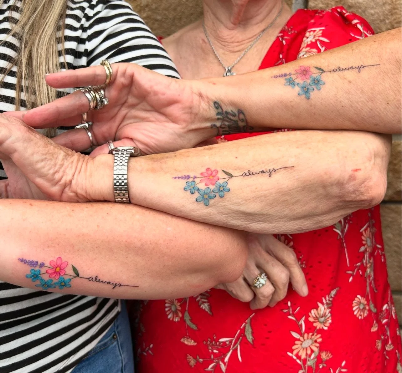 Multiple people with tattoos of flowers and the word 'always' on their arms, wearing jewelry, and dressed in patterned clothing.