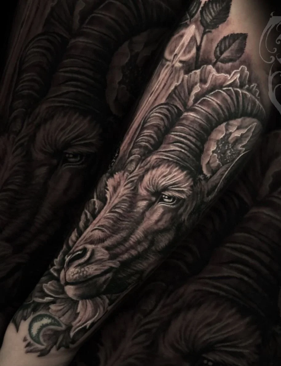 A close-up of a tattoo featuring a realistic ram with large, curved horns, surrounded by floral elements and intricate shading.