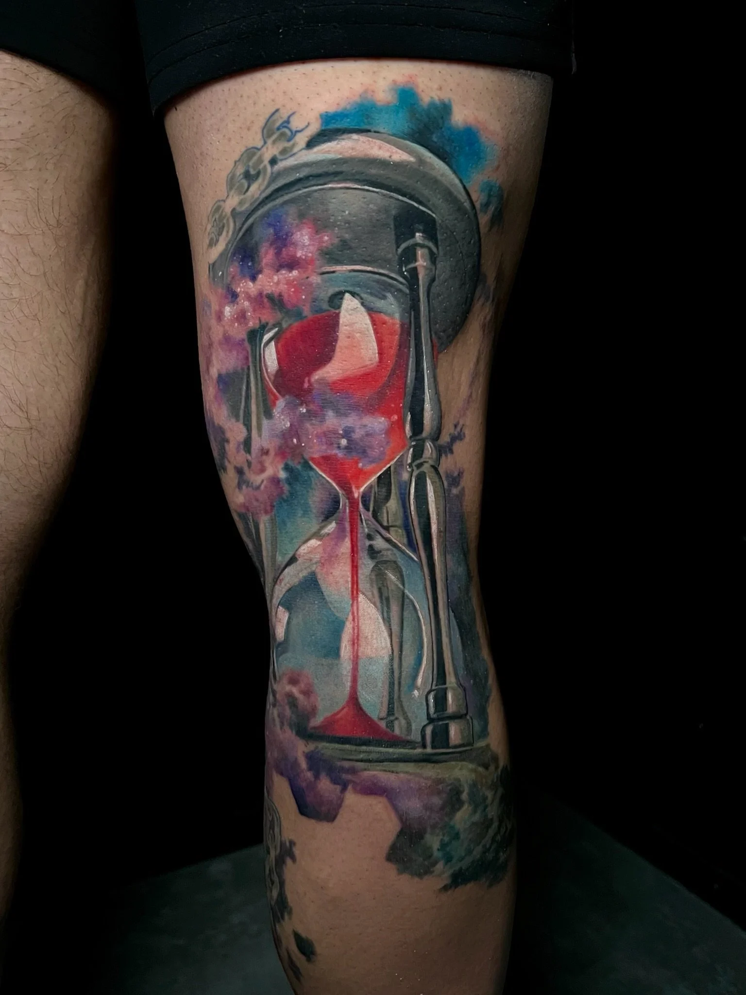 Tattoo of an hourglass with red sand near the bottom, surrounded by clouds and a city skyline at the top, with colorful clouds and sky in the background.