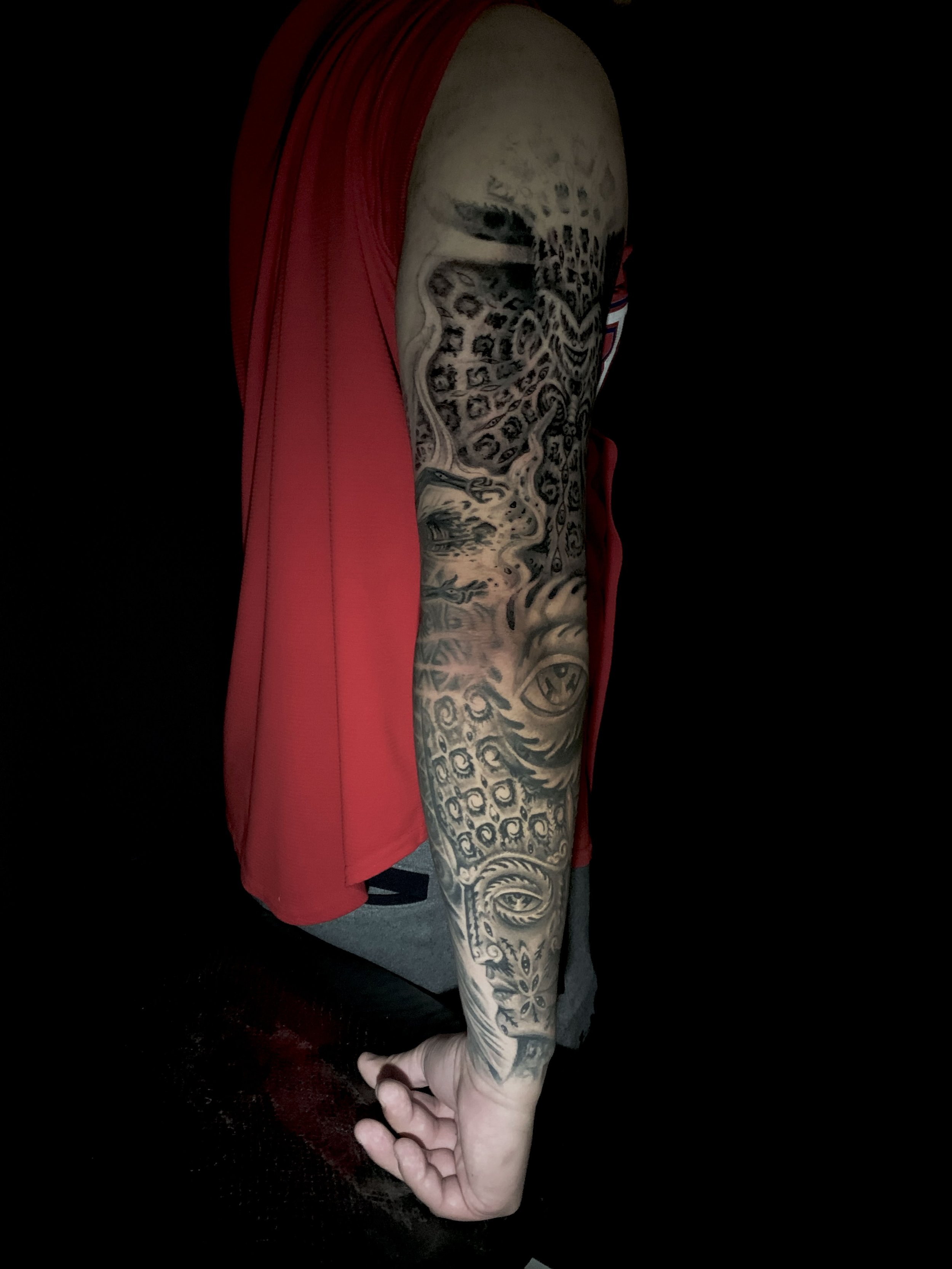 A tattooed arm in a red sleeveless shirt and gray shorts, with a hand extended downward, against a black background.