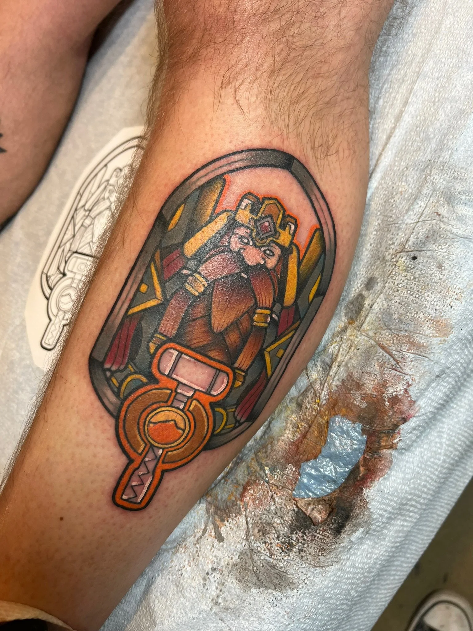 Tattoo of a king with a crown and beard, dressed in armor, inside a stained glass window with a hammer at the bottom.