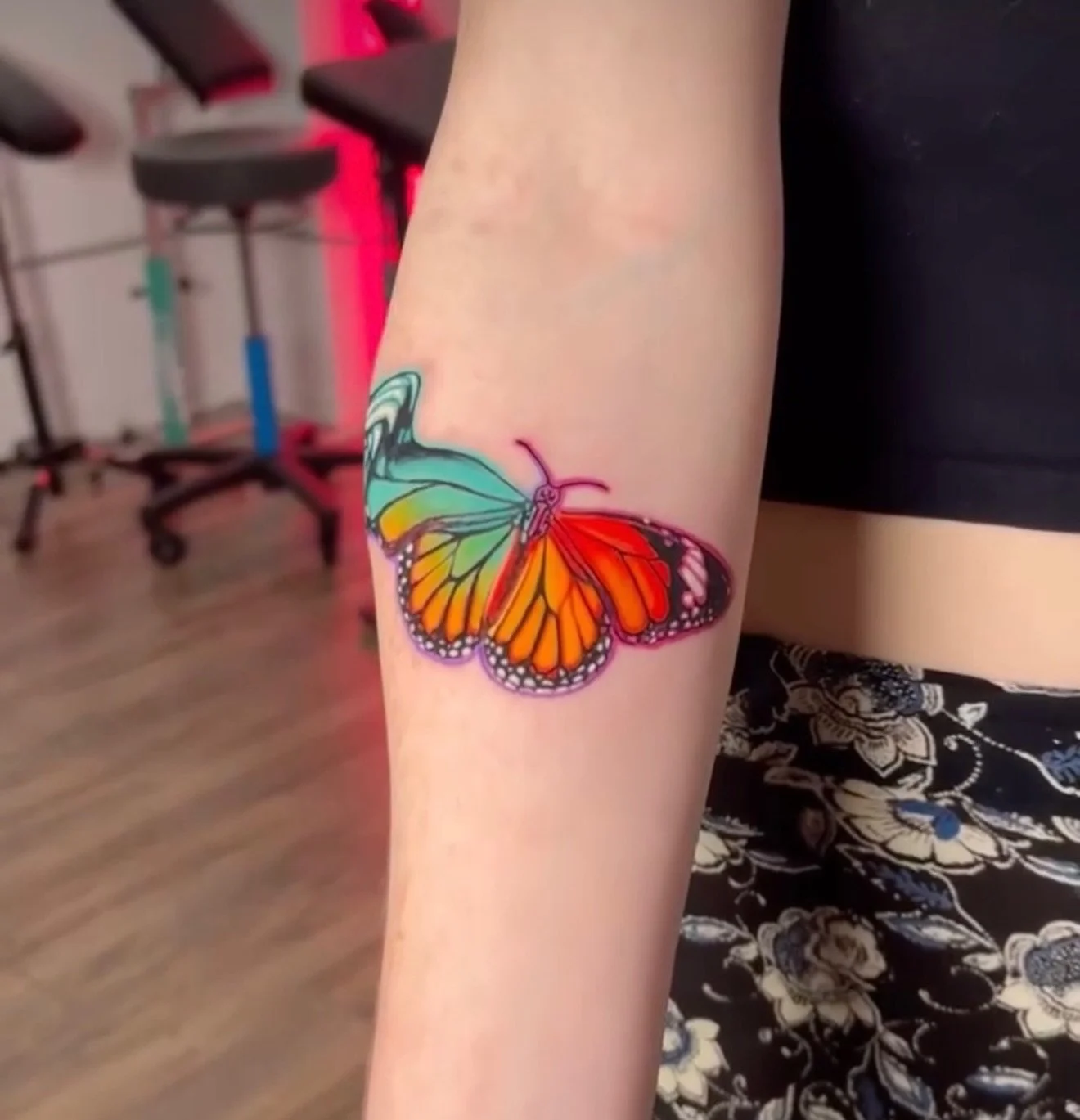 Colorful butterfly tattoo on person's forearm, showing orange, yellow, green, and black wings.