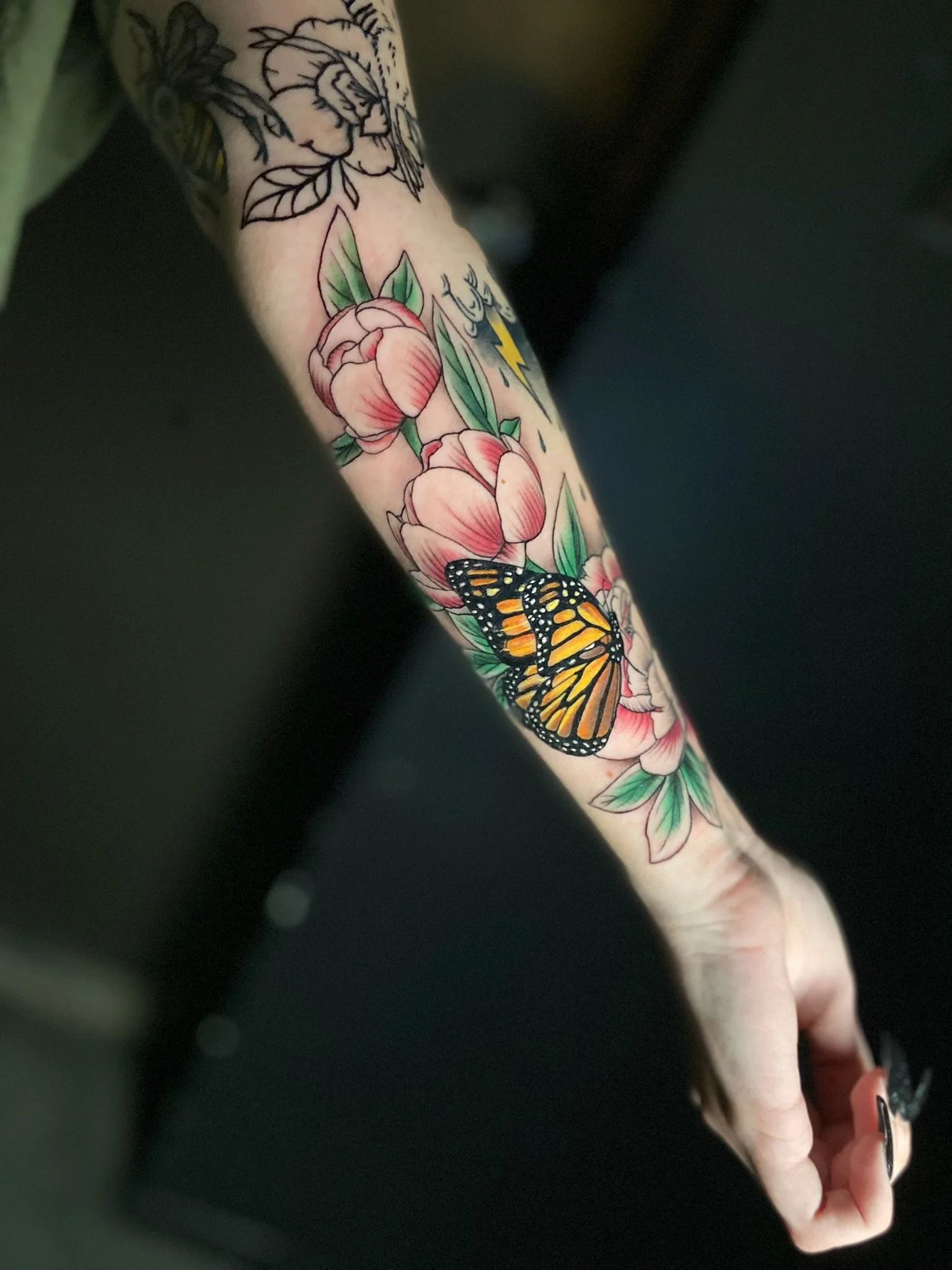 Colorful tattoo on arm featuring pink peony flowers, a Monarch butterfly, a lightning bolt, and water droplets.