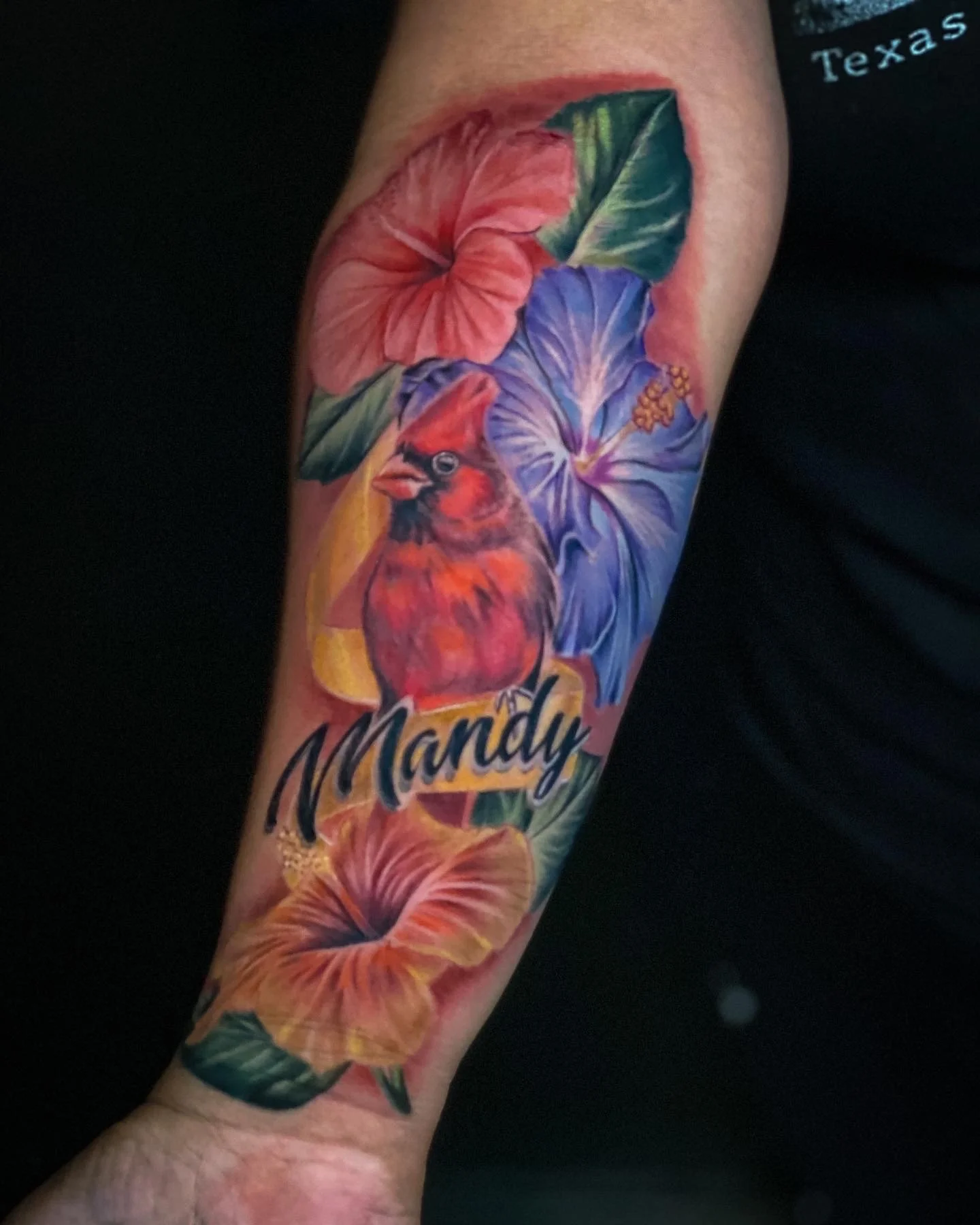 Colorful tattoo of a Robin bird, hibiscus flowers, and tropical leaves with the name 'Mandy' on the person's forearm.