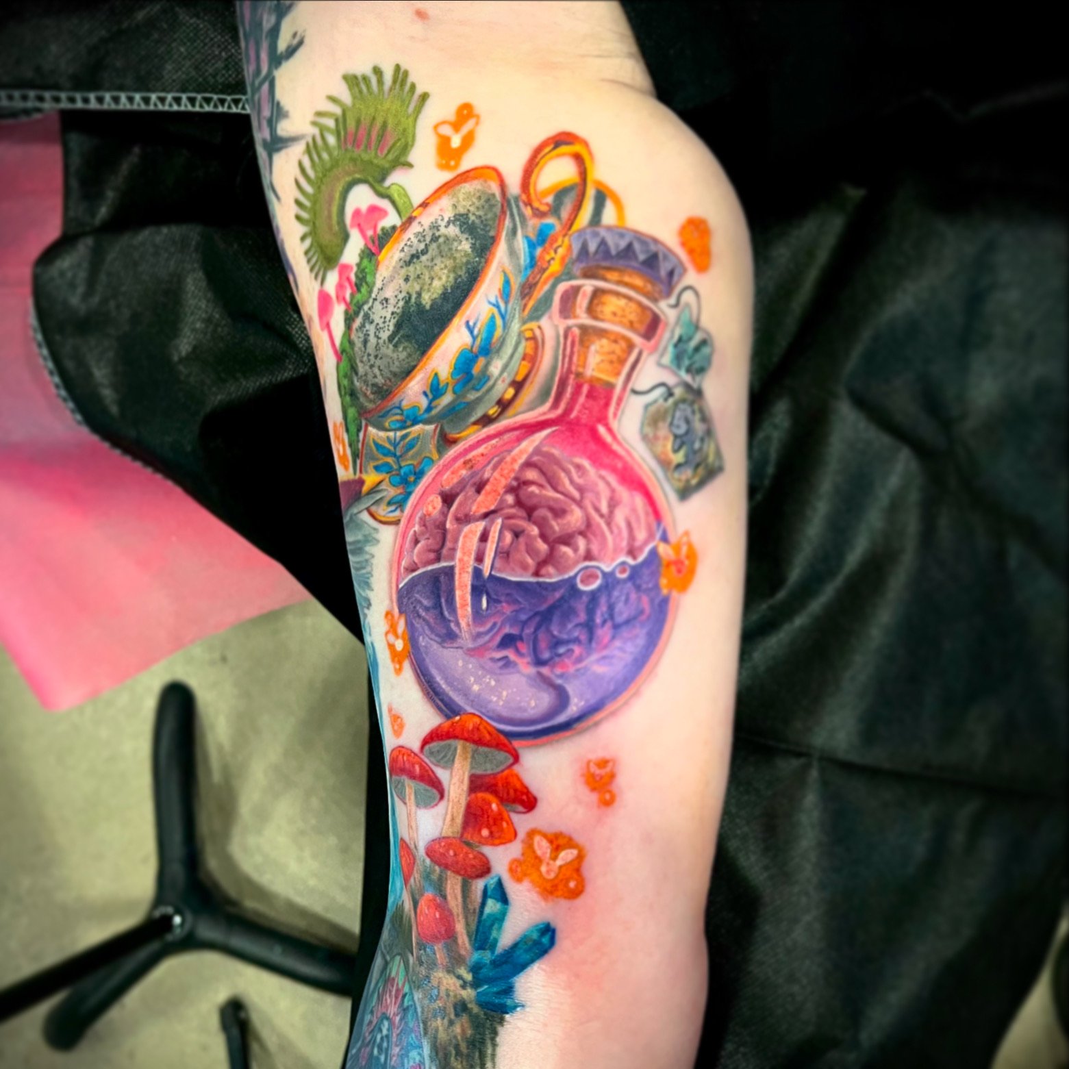 Colorful tattoo featuring a potion bottle with a brain inside, mushrooms, butterflies, a cup, and landscape elements.