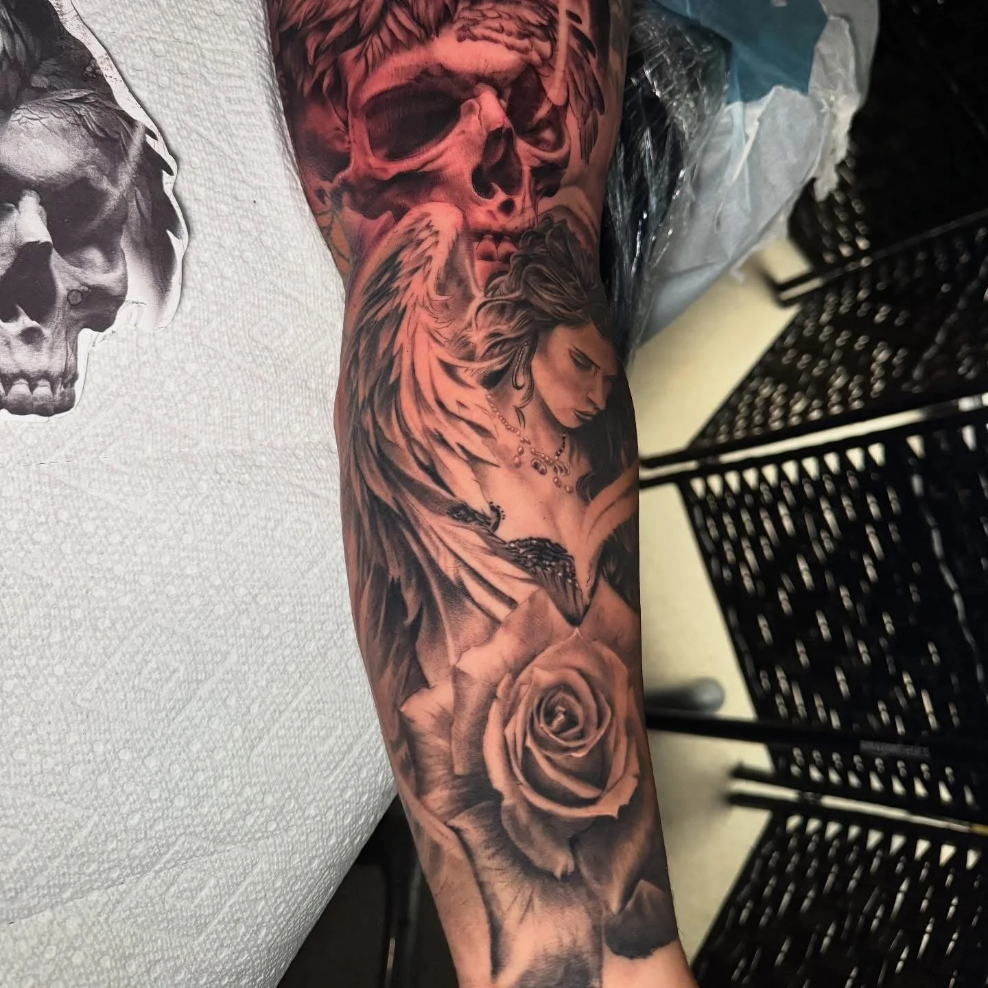 A detailed black and gray tattoo of a woman with long hair, a necklace, and a tattoo of a snake on her arm, surrounded by roses and a large skull with wings on the upper arm.