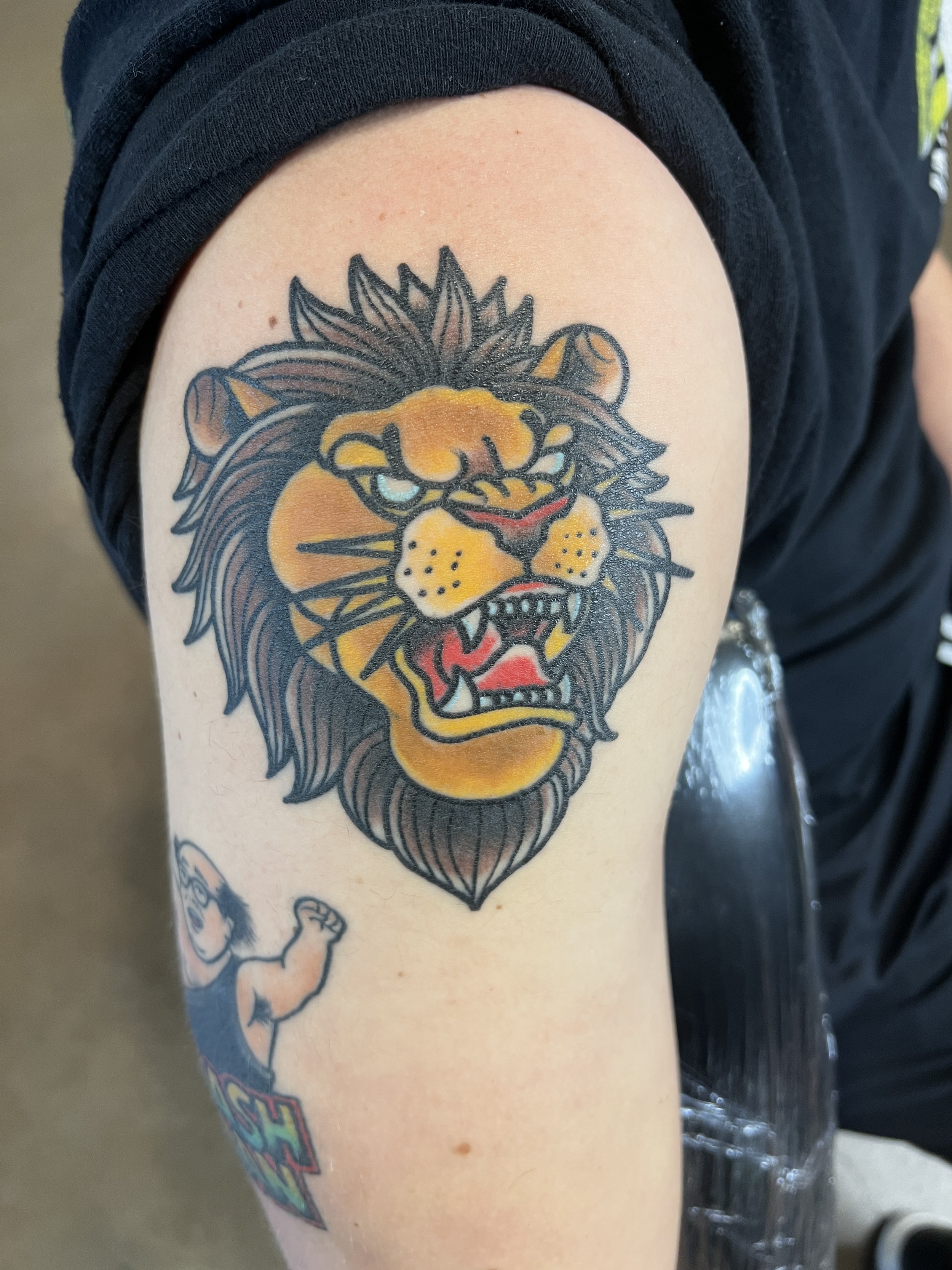 A tattoo of a fierce lion's face on a person's upper arm. The lion has blue eyes, a detailed mane, and an open mouth showing sharp teeth and a red tongue.