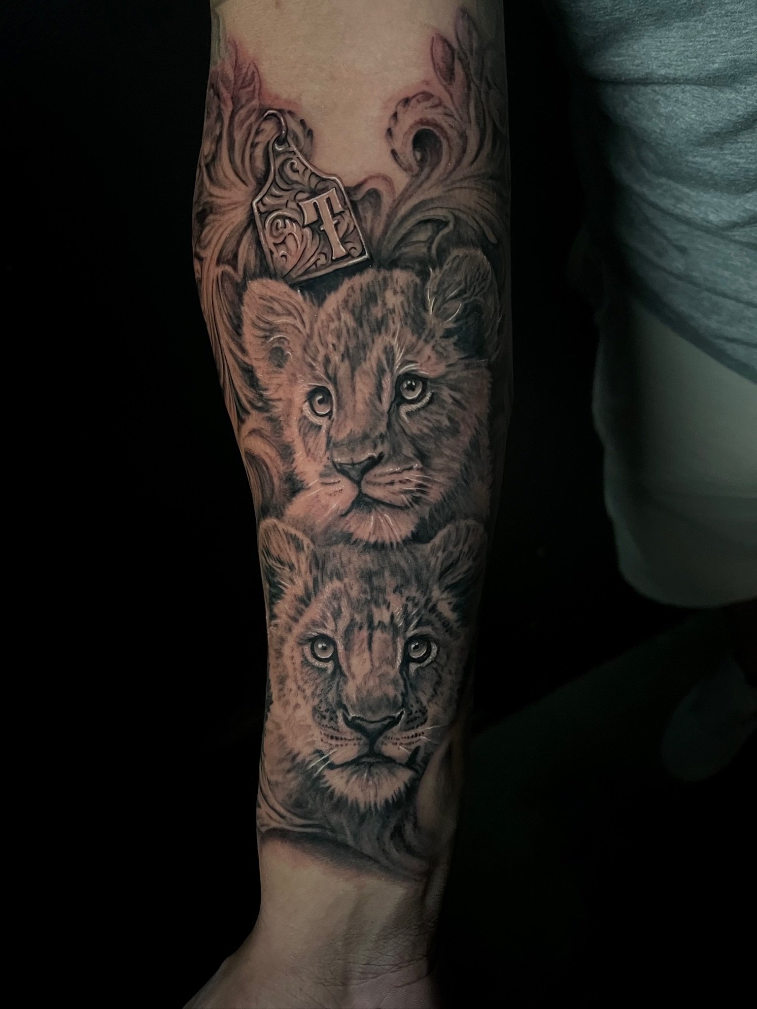 Tattoo of two lion cub faces stacked vertically on forearm, with a small crown and decorative swirl patterns at the top.