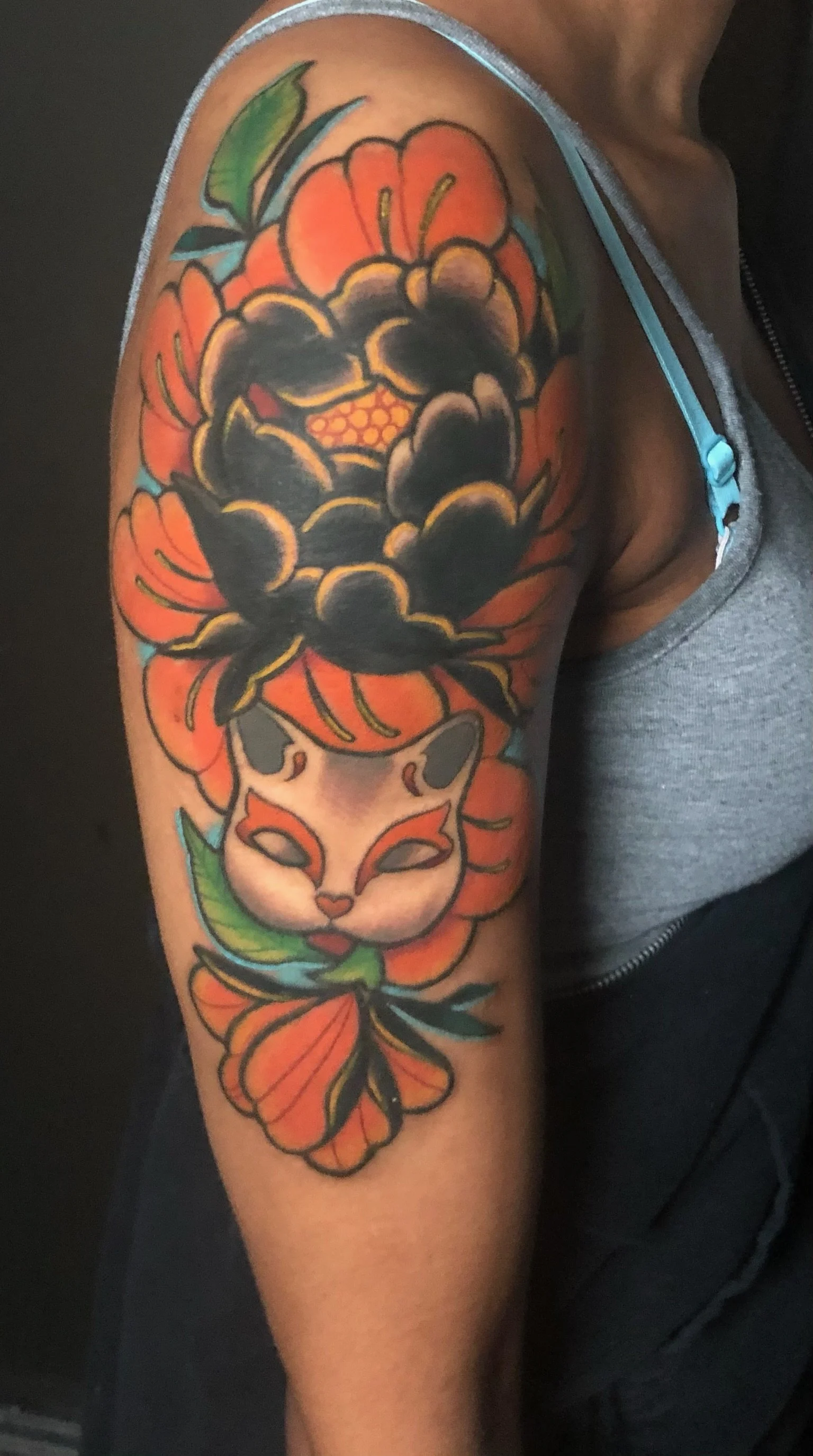 Colorful tattoo of a fox face with orange, white, and black fur, surrounded by large orange and black flowers, with green leaves, on a person's upper arm.