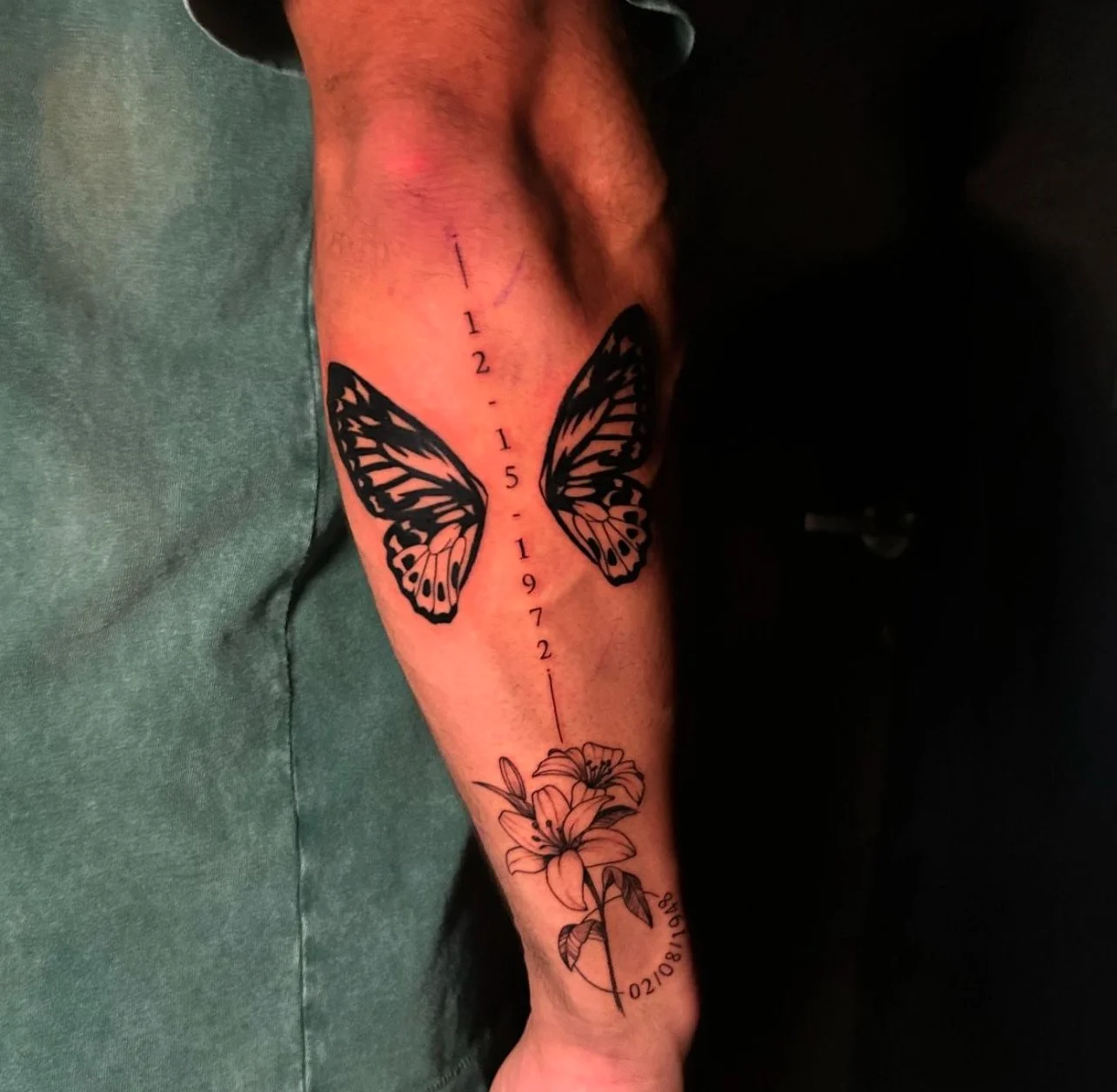 Tattoo of a butterfly with black and white wings, a vertical date with numbers 1, 2, 15, 1972, and a lily flower at the bottom on person's arm.