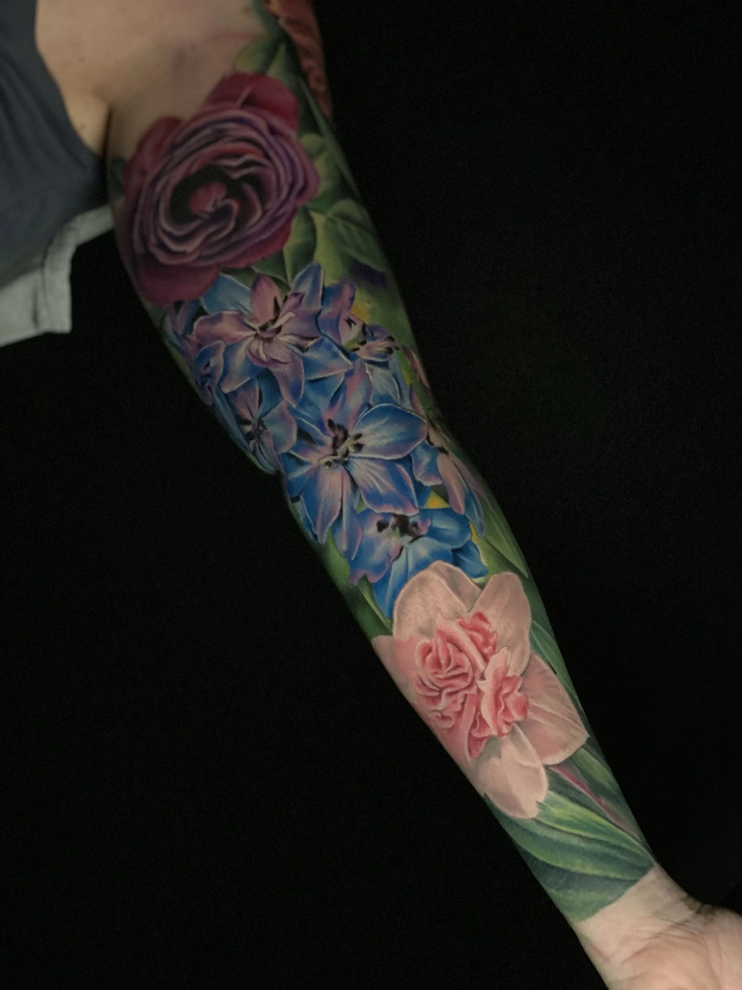A forearm tattoo of colorful flowers, including a pink rose at the wrist, purple and pink lilies, and additional flowers and leaves extending up the arm.