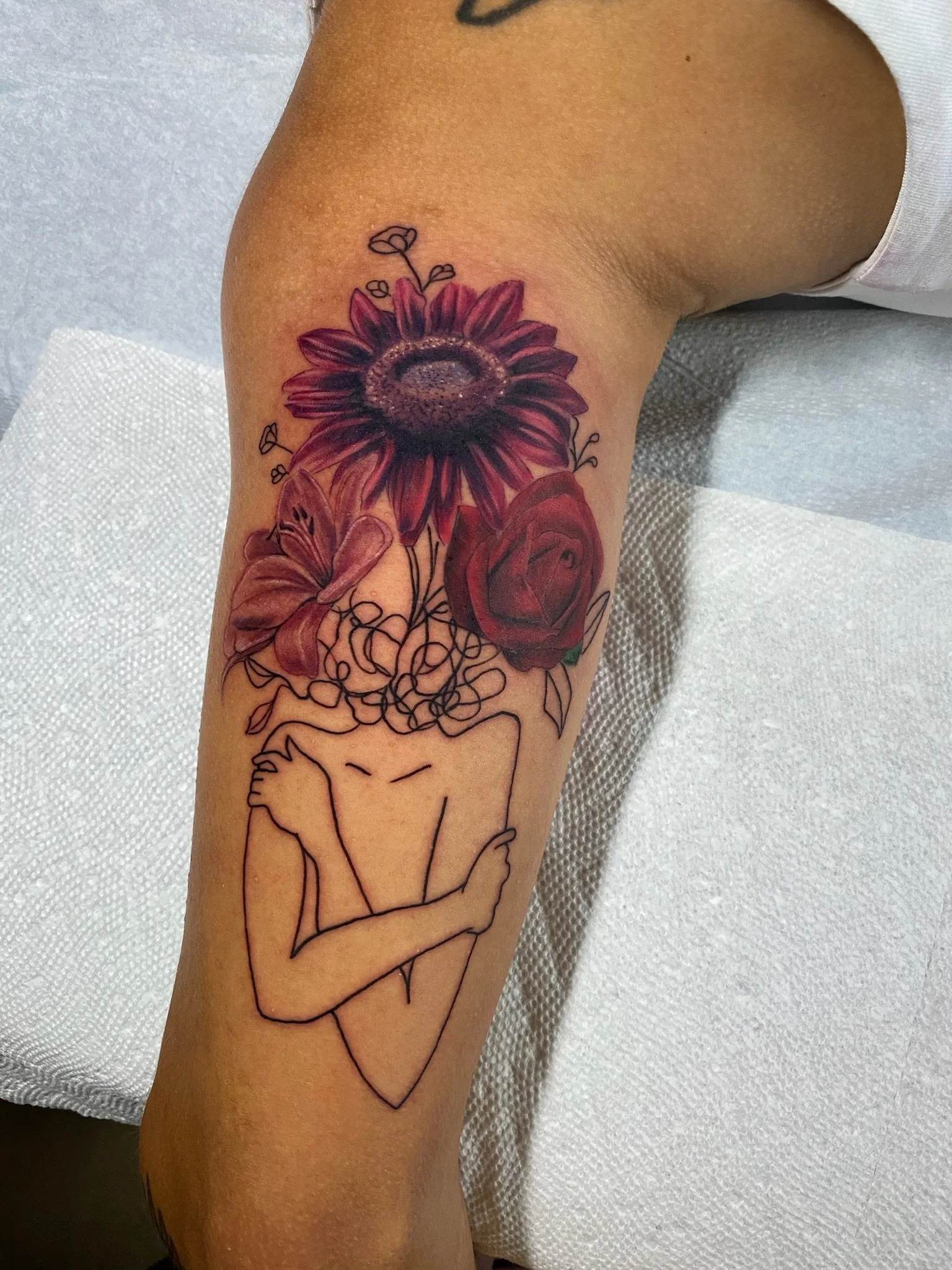 A tattoo on a person's arm featuring a colorful sunflower, a red rose, and a purple flower, with a line drawing of a woman with crossed arms below the flowers.
