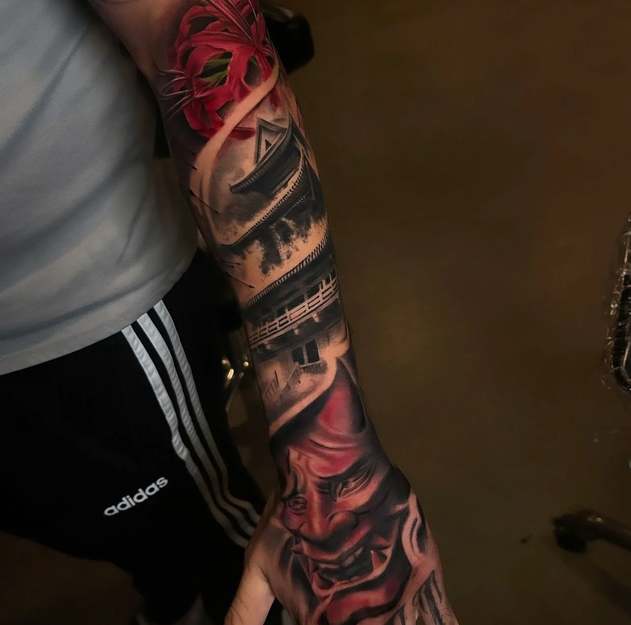 Close-up of a person's arm with a colorful tattoo sleeve, including a clown face, buildings, and abstract designs, while wearing a gray shirt and black Adidas shorts.