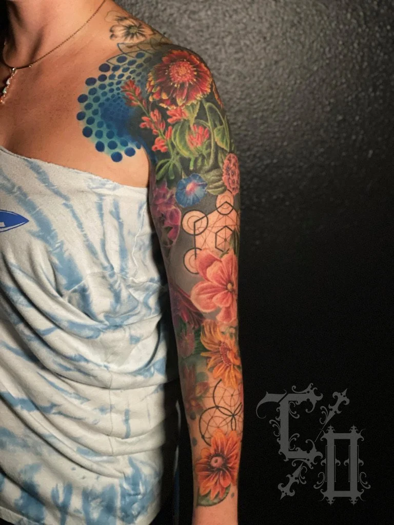 Colorful full sleeve tattoo of flowers, a bird, geometric patterns, and abstract elements on a person's arm and shoulder.