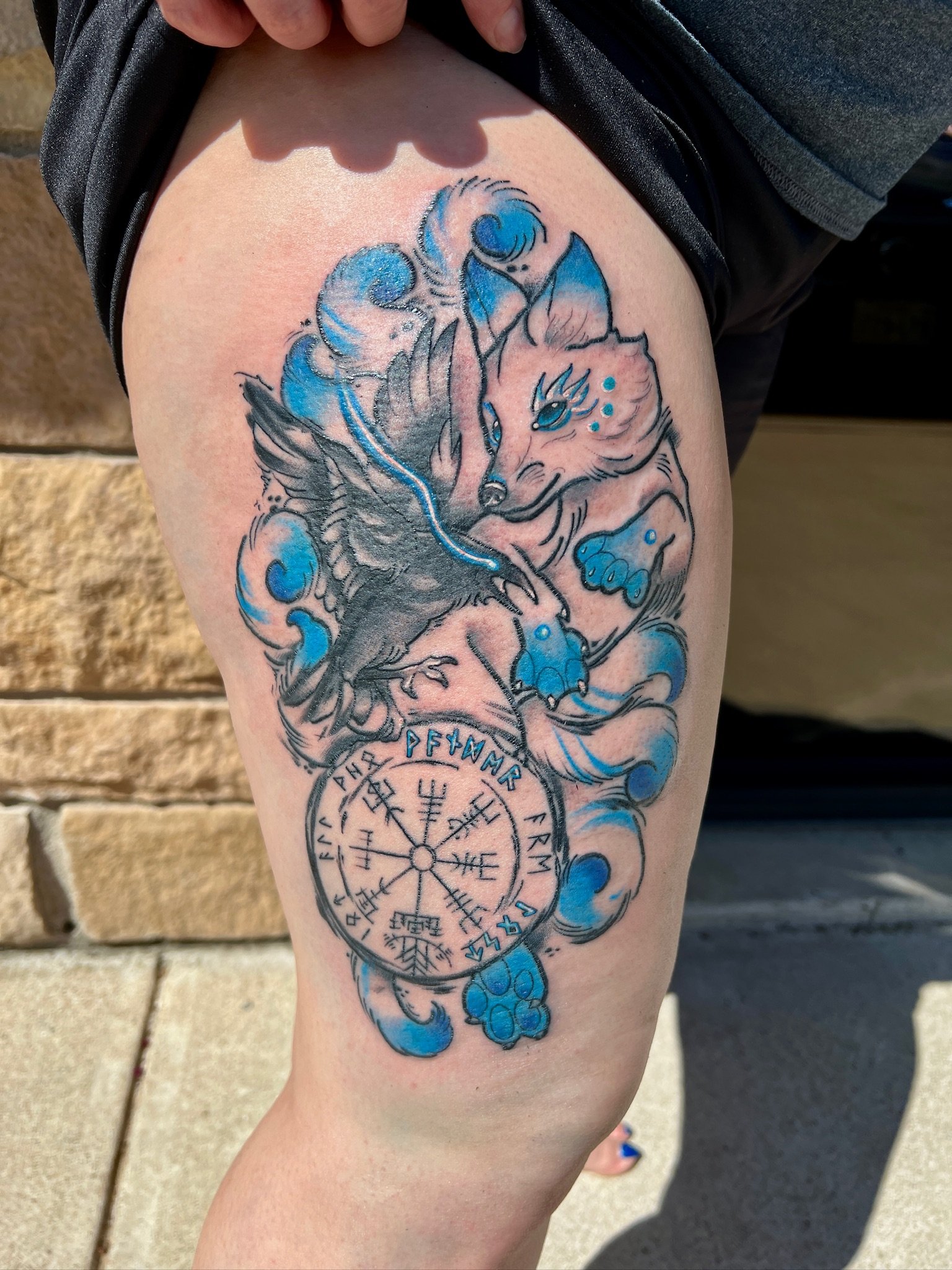 Tattoo of a wolf and a bird with a Norse rune circle and blue accents on a person's thigh.